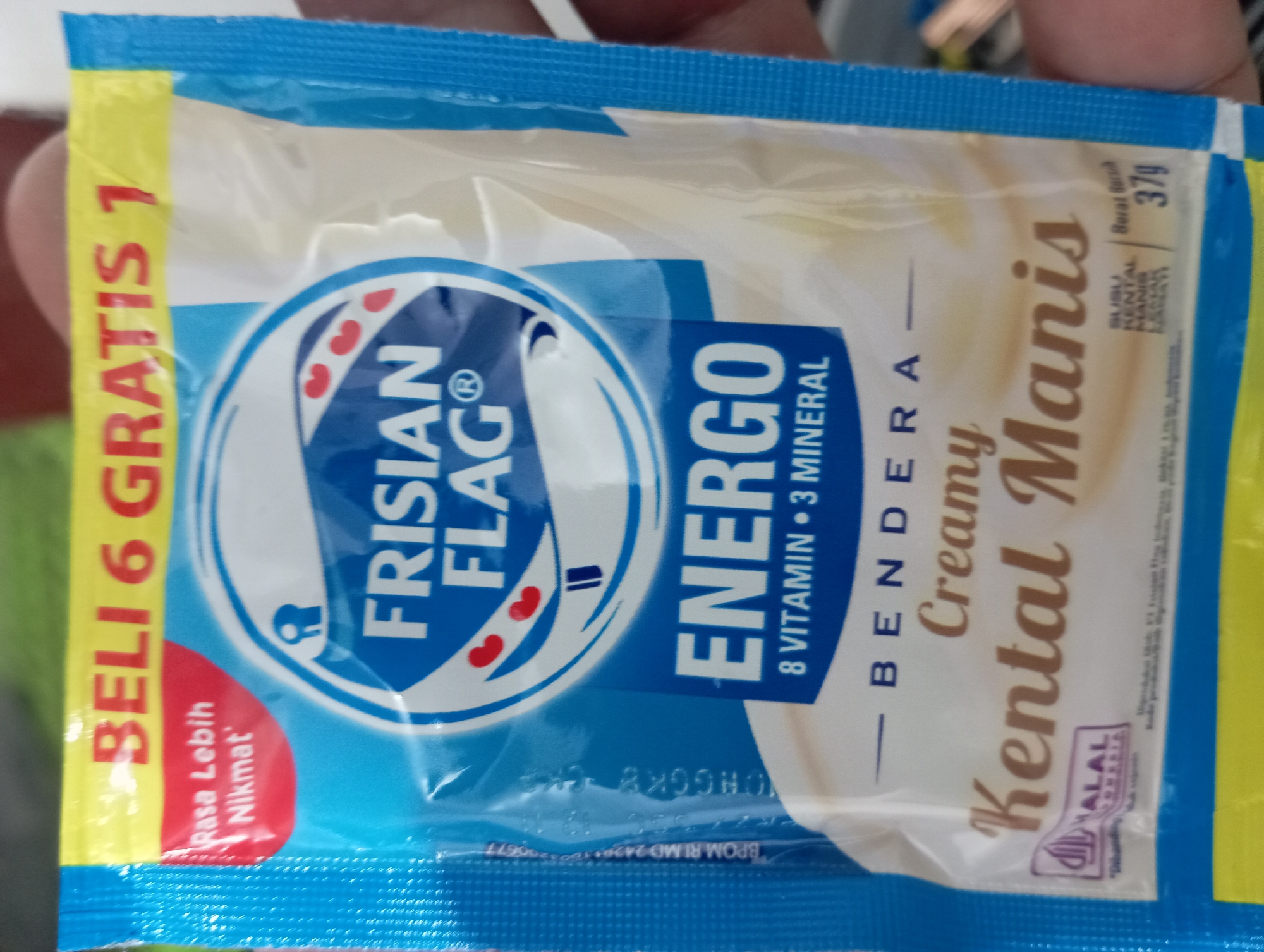 The image is a close-up shot of a Frisian Flag Energo creamy condensed milk packet. The packet is blue with yellow and red accents. Text on the packet includes "BELI 6 GRATIS 1" (Buy 6 Get 1 Free), "Rasa Lebih Nikmat" (More Delicious Taste), "FRISIAN FLAG", "ENERGO 8 VITAMIN 3 MINERAL", "BENDERA", "Creamy", and "Kental Manis" (Condensed Milk). A Halal logo for Indonesia is visible on the lower left. The net weight is indicated as "37g". The packet appears to be in good condition, with no visible damage. The lighting suggests it is indoors or in shade. There is no indication of the city of Kedungwuluh, Indonesia in the image.