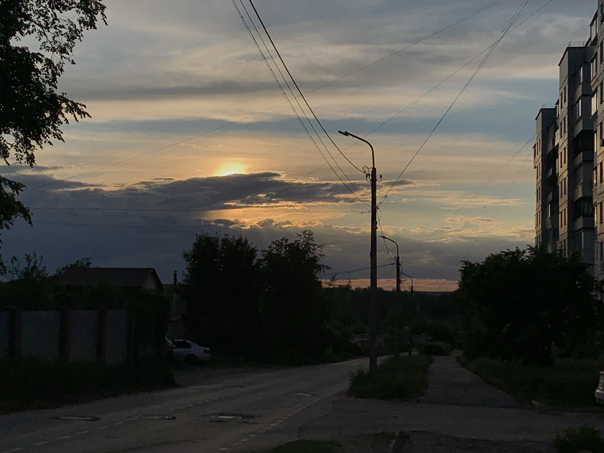 A landscape photo taken in the city of Chaikovsky, Russia. The time appears to be sunset. The photo is taken from a street showing a building on the right side. The street has street lights and telephone poles along it. On the left of the street is a house behind a fence. A vehicle is partially visible behind the fence. There is a tree on the left of the image. The sky has clouds and the sun is visible through them. The overall tone is dark.