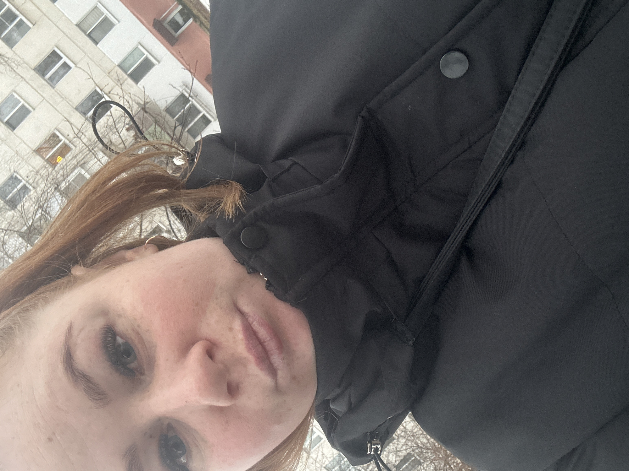 A close-up, slightly angled, selfie photograph shows a fair-skinned person with reddish-brown hair and freckles. They are wearing a black winter coat with buttons. Behind the person, an apartment building is visible, suggesting an urban location. The presence of bare tree branches implies it is either late fall, winter, or early spring. There is not enough information to accurately pinpoint the exact location within the city of Chaikovsky, Russia.
