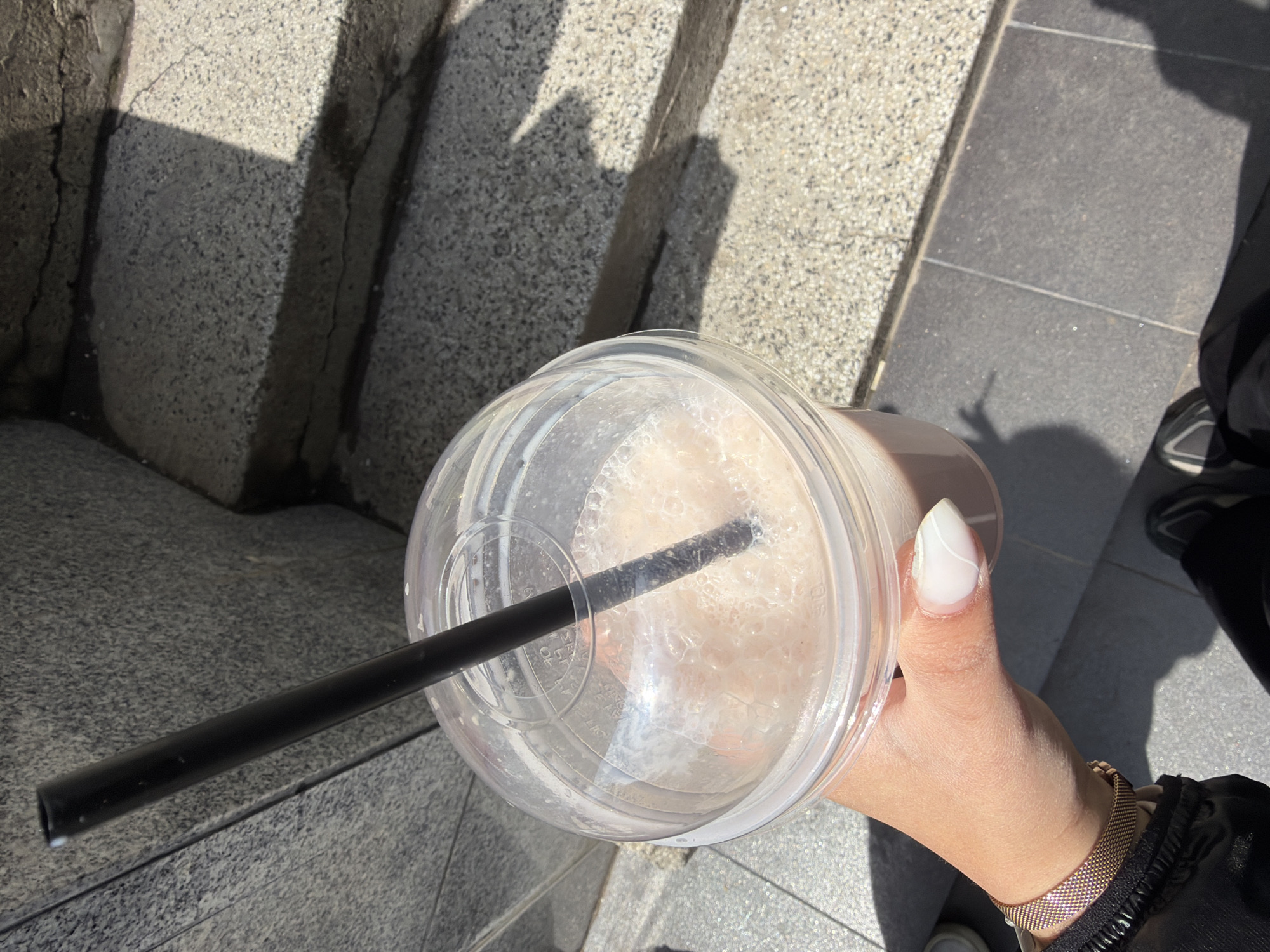 A high-angle, outdoor shot captures a person's hand holding a large, clear plastic cup containing a light-colored, foamy beverage, likely a milkshake or iced coffee. A black straw protrudes from the plastic domed lid. The person's right hand features manicured, long, oval-shaped nails with a white tip or full white polish, and a subtle white line detail on the thumb. A gold-toned mesh watch band adorns the wrist, and part of a dark garment (possibly a jacket sleeve) is visible.
The scene is set outdoors in Sofia, Bulgaria, on a paved surface. The ground consists of textured, speckled grey stone or concrete, alongside smoother, darker grey rectangular paving tiles. Strong, direct sunlight casts prominent, angular shadows across the ground and adjacent steps or ledges, indicating a bright, clear day, likely during the morning or late afternoon. Faint, molded text, including characters like "DIT" and numbers, can be observed on the plastic lid of the cup. The image conveys a casual moment of refreshment on a sunny day.