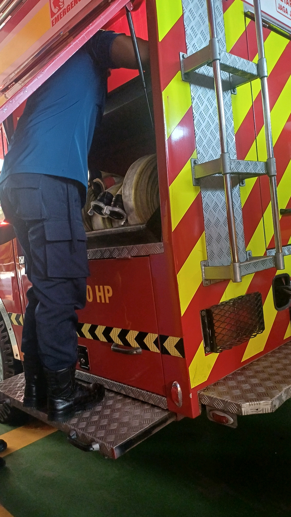 In South Jakarta, Indonesia, a firefighter in navy blue pants and a blue shirt is seen stepping onto the deployed metal platform of a red and yellow striped fire truck. The firefighter is looking down into the truck, where several coiled hoses are visible. The truck has the text "250 HP" printed on its side in yellow lettering. The scene appears to be indoors, possibly in a fire station, given the presence of a green floor and the controlled lighting.