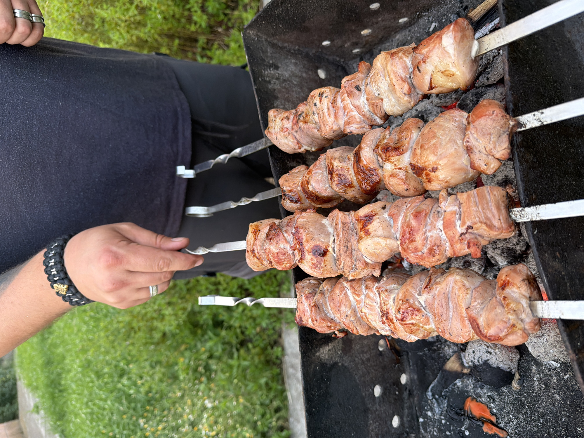 A person is cooking meat skewers over a charcoal grill outdoors. The meat is cubed and skewered onto metal rods, with several skewers already placed on the grill and others being adjusted by the person's hand. The grill is filled with hot coals, some of which are glowing red. In the background, green foliage and grass are visible, suggesting a natural or park-like setting. The person is wearing a dark t-shirt and has rings on their fingers and a bracelet on their wrist. The overall scene depicts a casual outdoor cooking activity. The location is identified as Lozuvatka, Ukraine.