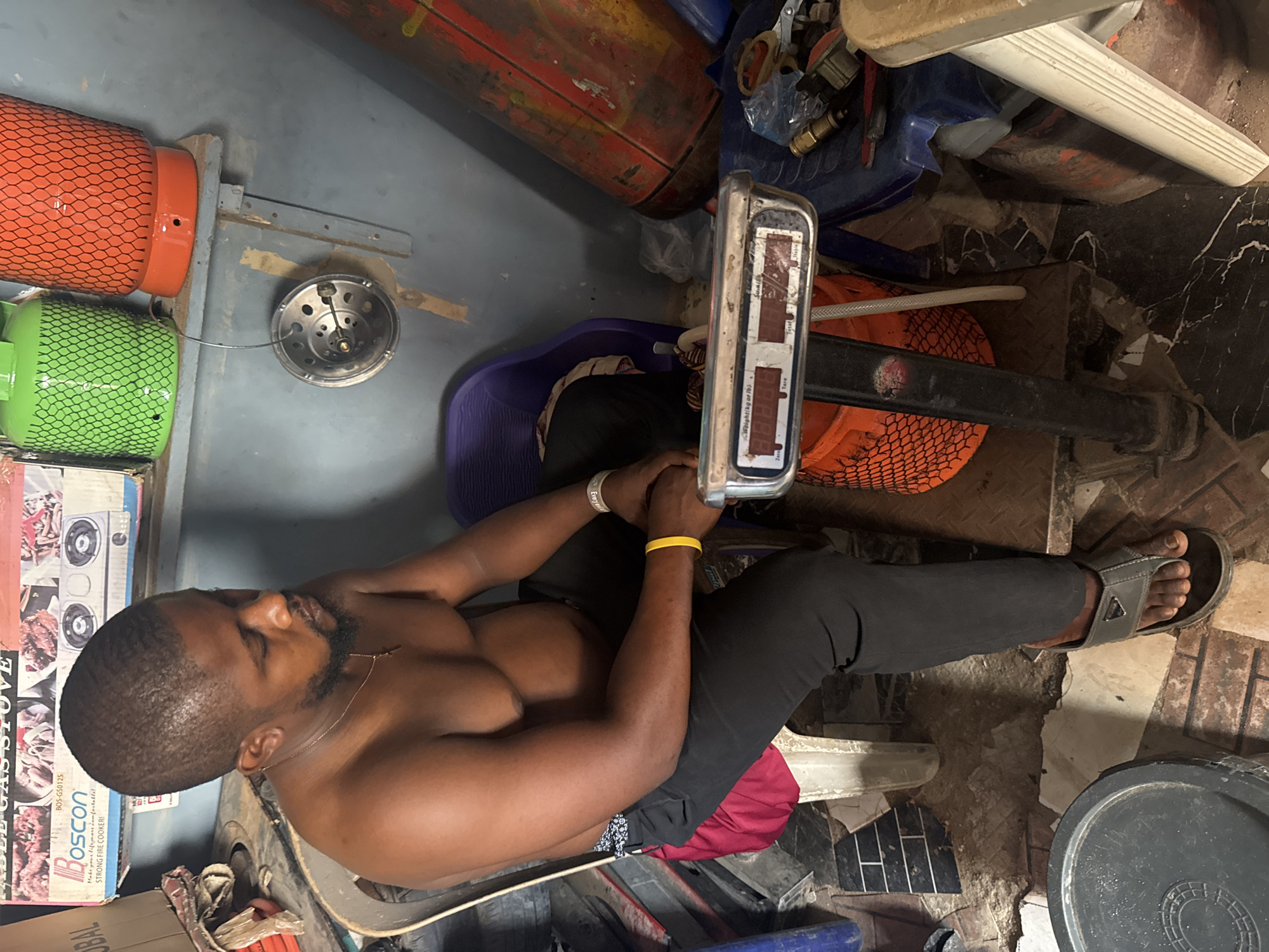The image depicts a man engaged in an activity likely related to gas cylinder sales or refills in Ihiagwa, Nigeria. The man, who is shirtless and wearing dark pants and flip-flops, is seated and has his hands resting on a digital weighing scale. He has short hair, a beard, and wears both a yellow and a white wristband (the latter partially legible as "Everyth"), along with a thin necklace.
The scene is cluttered with various objects, most notably several gas cylinders. A prominent green and an orange gas cylinder, both enclosed in mesh, are visible on the left, with a hose connecting to a circular metal gas stove burner. Other red or orange gas cylinders are scattered in the background. The digital scale shows indicators for "Weight (kg or lb)", "Total", "Tare", and "Zero".
To the left, a cardboard box is visible with text including "BOS-G50125", "Boscon", and "STRONG FIRE COOKER", along with the slogan "Make your life more comfortable!". The floor consists of concrete interspersed with sections of broken or uneven tiles. The overall setting appears to be a functional, possibly commercial, space with various tools and containers around. The man's focused gaze downwards suggests he is concentrating on the task at hand, likely weighing the gas cylinders. The lighting indicates it is daytime.