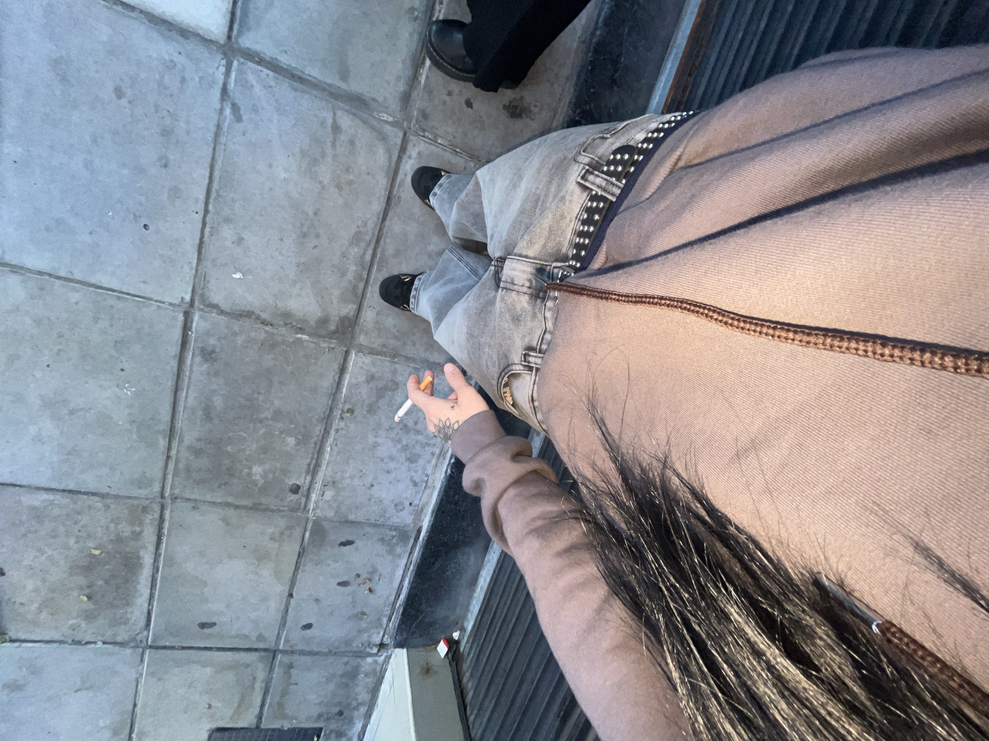 A high-angle shot captures a person standing on a grey tiled sidewalk, holding a lit cigarette in their left hand. The person is wearing light wash, wide-leg jeans with a prominent black studded belt, and a light brown or beige hooded top. Their long dark hair is visible falling over their shoulder and arm. A tattoo of a delicate design is visible on the back of their left hand. They are wearing black shoes, possibly loafers or chunky flats. In the background, partially visible, are the black shoes and lower legs of another person, standing on the same tiled surface. The sidewalk appears somewhat weathered with visible grout lines and some stains or dirt. To the right of the primary subject, a dark, possibly metallic or slatted structure, like a fence or wall, runs parallel to the ground. The lighting suggests it is daytime, possibly overcast. No specific text is visible in the image. The scene depicts an urban outdoor setting, consistent with a sidewalk in Buenos Aires, Argentina.