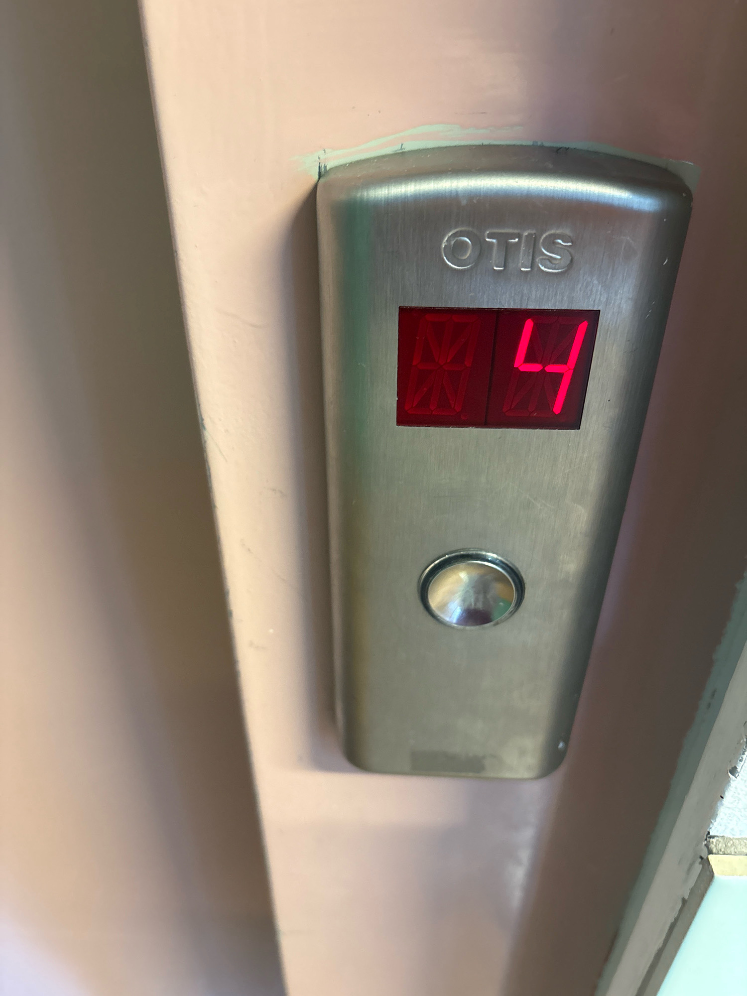 The image shows an Otis elevator panel with a digital display showing "04". The panel is made of brushed metal and is mounted on a light pink wall. There is a circular button below the display. The scene appears to be indoors, likely in a building in Reykjavik, Iceland, though there are no specific identifiers to confirm this location. The lighting is somewhat dim, with reflections visible on the metallic surfaces.