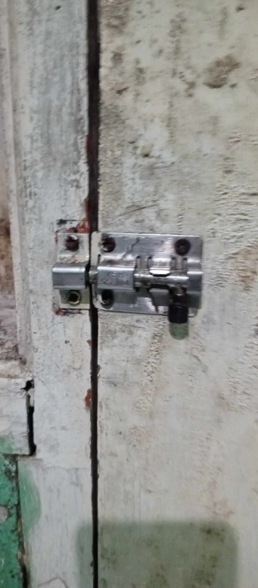 The image shows a close-up of a sliding bolt latch mechanism attached to a weathered, light-colored wooden door. The latch is made of metal and is secured with screws. The door appears old and worn, with visible cracks and stains. The background consists of the textured surface of the door and a dark vertical gap, suggesting two parts of the door are meeting. The lighting is dim, and the overall impression is one of decay and disuse. This scene is not specified to be in Karawang, Indonesia.