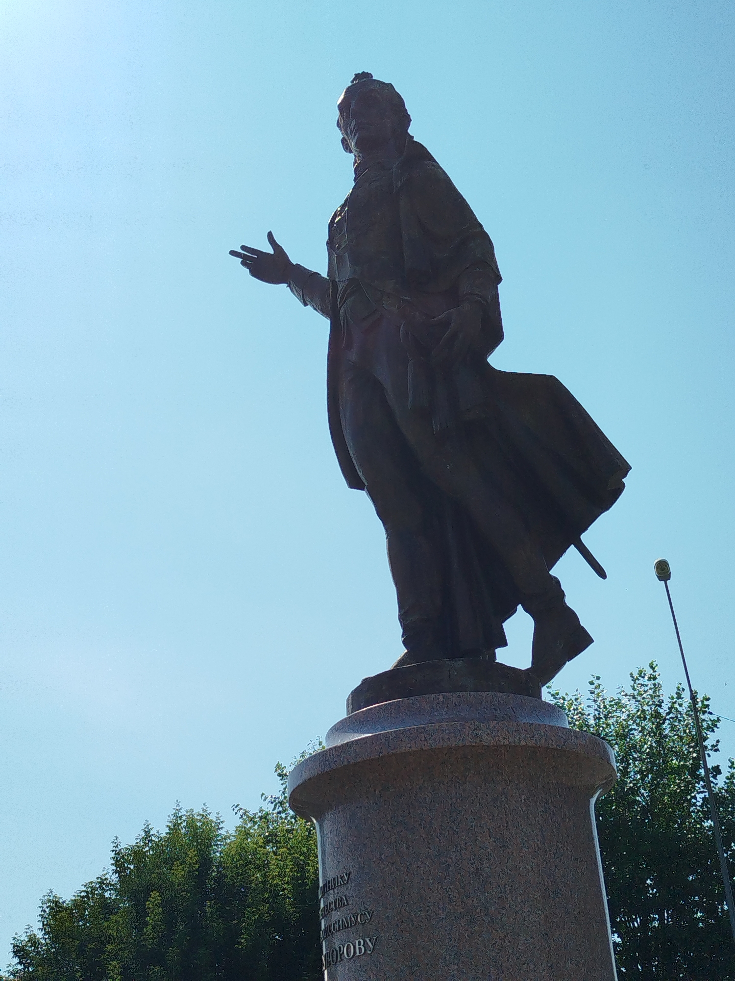 The image shows a bronze statue of a man in historical attire, standing on a stone pedestal. The statue is viewed from a low angle against a clear blue sky. The man is depicted with his right arm extended and his hand open, as if speaking or gesturing. He wears a long coat and what appears to be a sword at his side. In the background, there are green trees and a lamppost.
The inscription on the pedestal reads "ПАМЯТНИКУ ВЕЛИКОМУ СЫНУ ЗЕМЛИ ТЮМЕНСКОЙ АЛЕКСАНДРУ ФЕДОРОВИЧУ СУРКОВУ". This translates to "To the monument to the great son of the Tyumen land Alexander Fyodorovich Surkov".
This statue is located in the city of Yalutorovsk, Russia. Alexander Fyodorovich Surkov (1907-1981) was a Soviet poet, playwright, and writer.