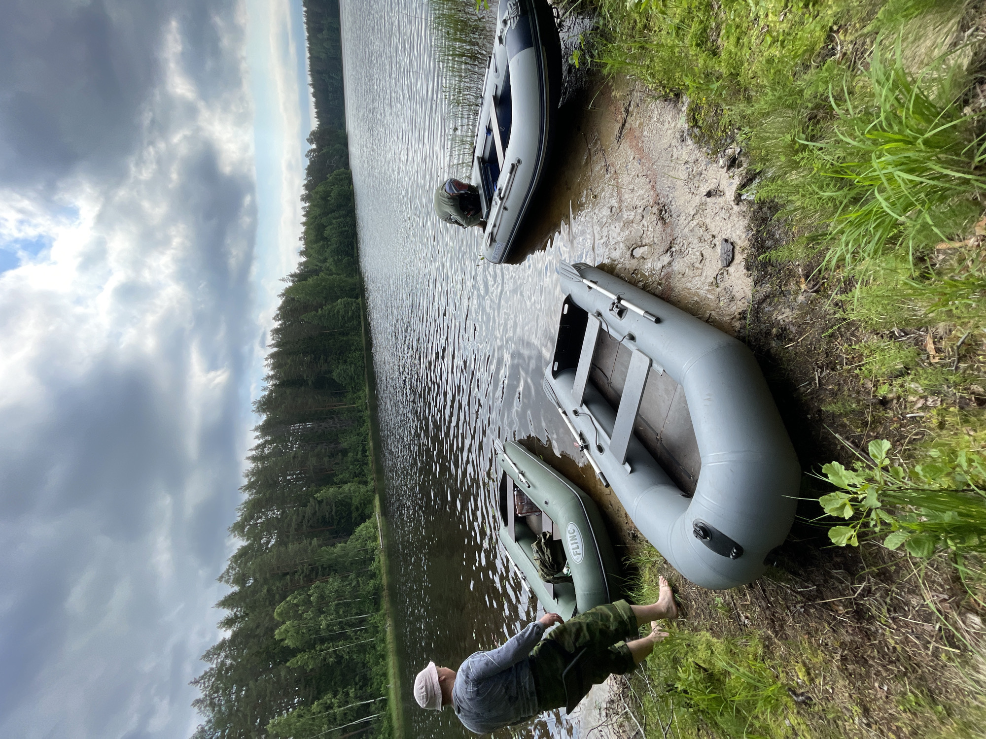A man wearing a light-colored hat, long-sleeved gray shirt, and camouflage pants stands on the muddy shore of a lake in Roshchino, Russia. He is barefoot and facing away from the camera, looking out at the water. Three inflatable boats are partially submerged in the lake, resting on the shore. The leftmost boat is green and has the brand name "FLING" visible. The other two boats are gray and appear to be larger. A person wearing a dark jacket is partially visible inside the gray boat furthest from the shore, appearing to be adjusting something near the outboard motor. The lake is surrounded by a dense forest of pine and deciduous trees. The sky is overcast with dark, dramatic clouds, suggesting a storm may be approaching. The scene conveys a sense of preparation for or rest after a water activity in a natural, possibly remote, setting.