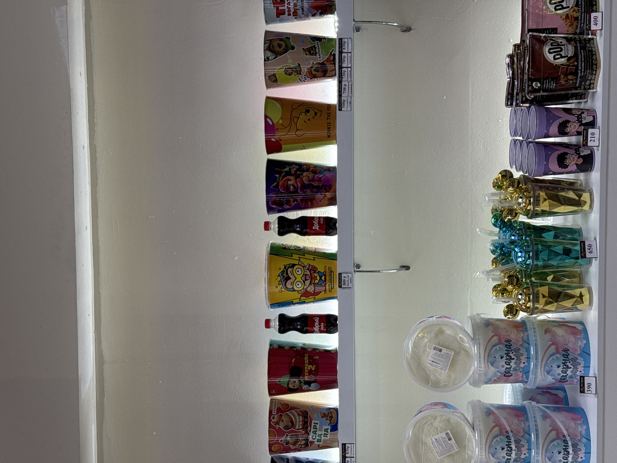 A vending machine in an indeterminate location in Unknown, Unknown is stocked with snacks and beverages. The machine features an illuminated interior that displays the items available for purchase. From top to bottom, the items appear to include popcorn, decorative cups with various cartoon character themes, cotton candy, and bottles of Coca-Cola. Prices are labeled next to each item.