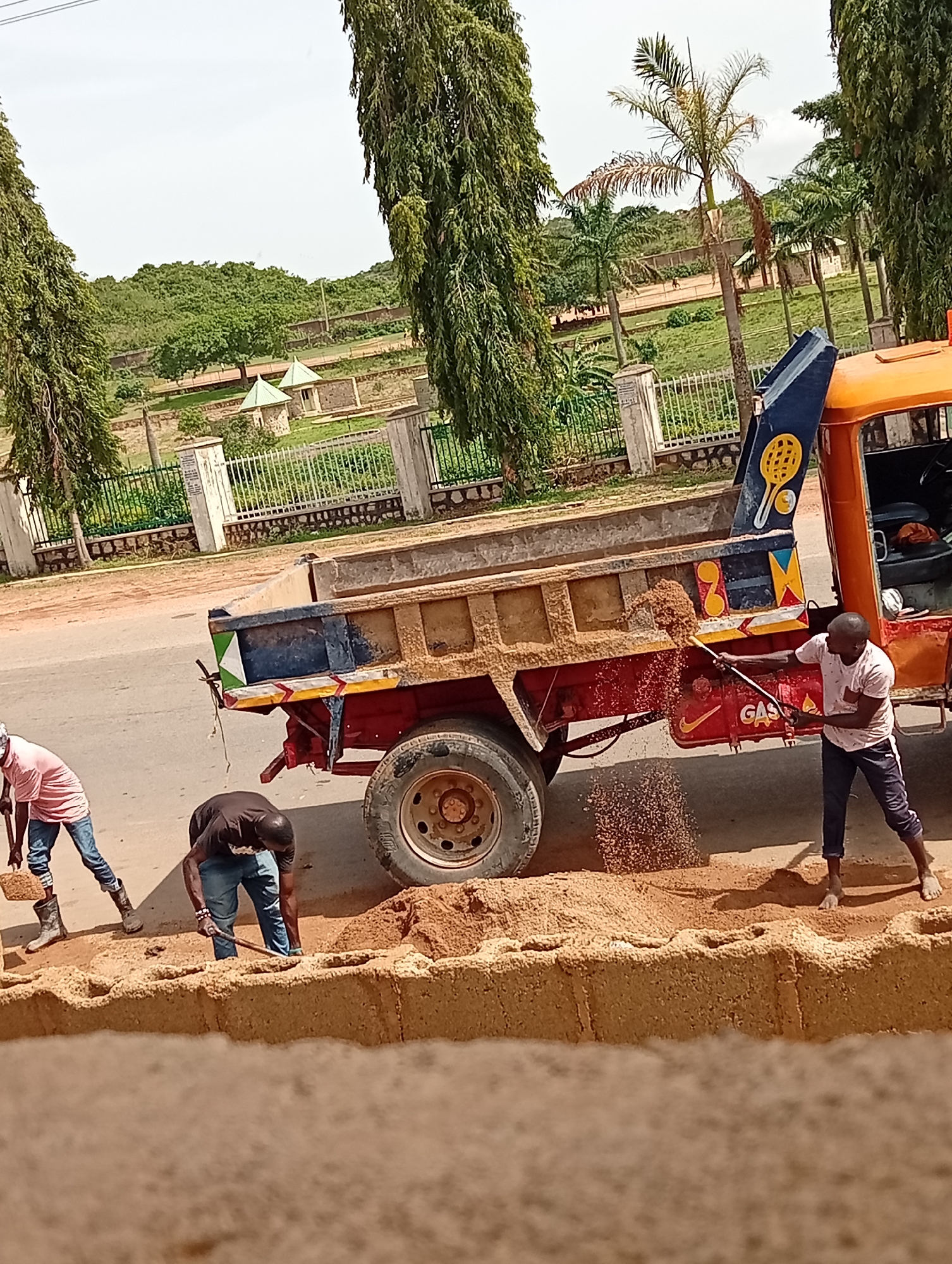 The image shows three men unloading sand from a dump truck in Jos, Nigeria. The men are using shovels to move the sand, which is forming a large pile on the ground. The truck is parked on the side of a road, with trees and buildings in the background. The area visible beyond the truck's bed is park-like, with trees and low buildings set behind a wrought-iron fence.