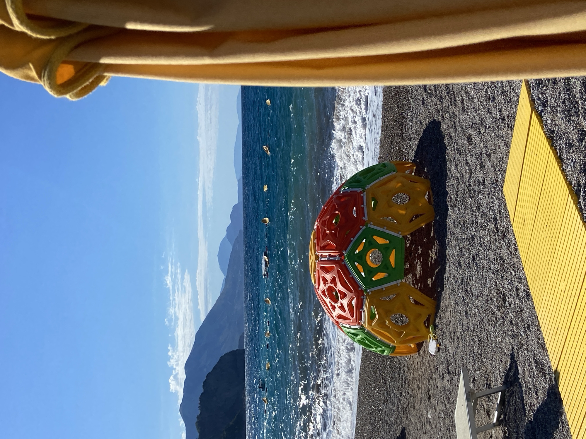 The image shows a beach scene in Prijevor, Montenegro, under a clear blue sky. In the foreground, a colorful, geodesic dome-shaped play structure, composed of red, green, and yellow plastic panels, sits on a pebble beach. To the left of the structure is a white lounge chair. In front of the structure and extending towards the right is a yellow, slatted walkway. A large yellow curtain occupies the right side of the frame, partially obscuring the view. The ocean is visible in the background, with moderate waves breaking on the shore. Further out, several small buoys or markers are scattered across the turquoise water. In the distance, a chain of dark, rugged mountains meets the horizon under a hazy sky. There are no people visible in the image.