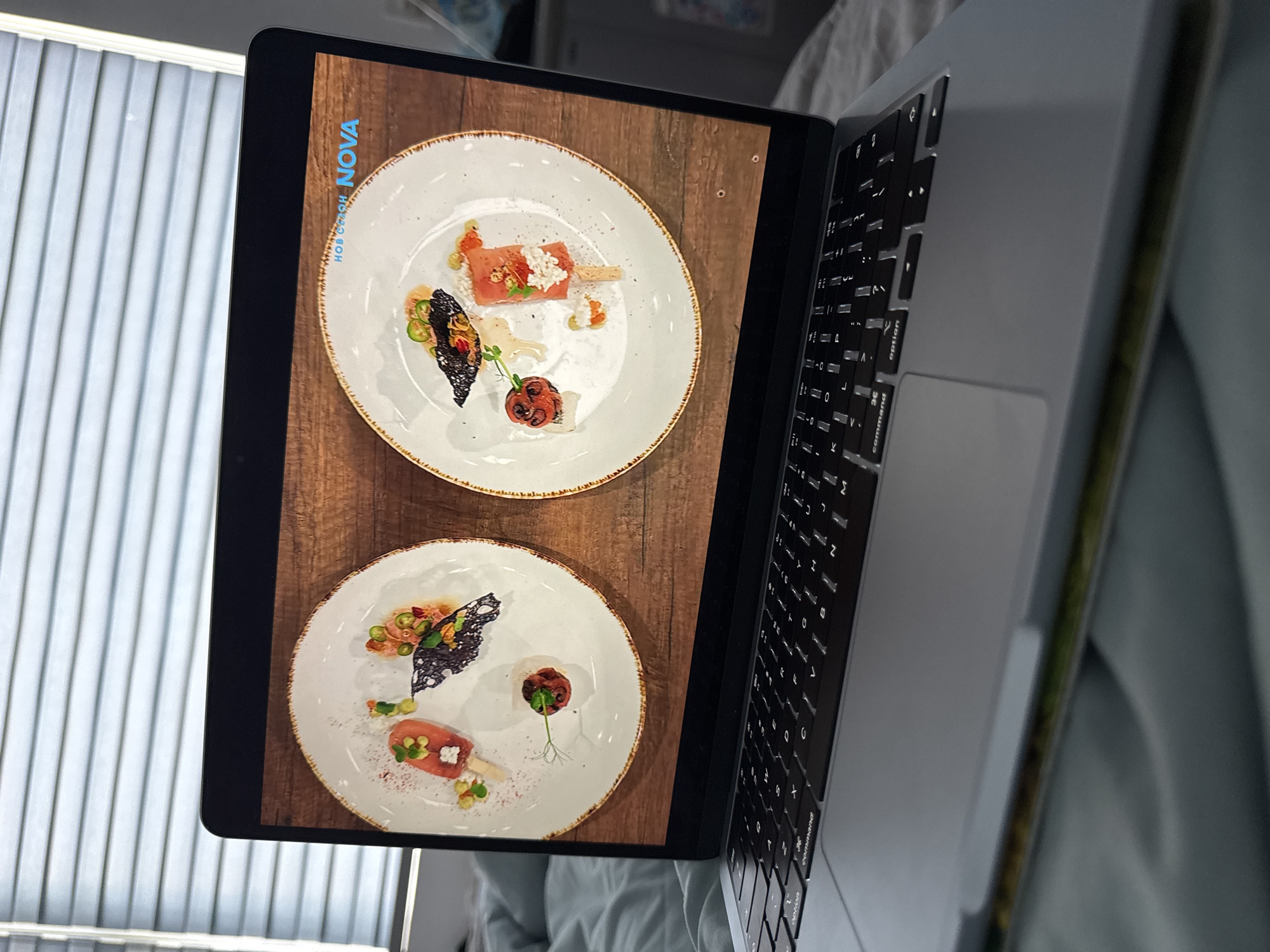 The image depicts a laptop screen displaying a photograph of two plates of food against a wooden background. Each plate contains a small, elaborately presented dish with a popsicle-like food item, garnish, and other small elements. The laptop is sitting on a soft, slightly wrinkled surface, possibly a bed or couch. The background includes a window with white blinds. There is no visible information to determine the location in Đà Nẵng, Vietnam, or any details about people or interactions.