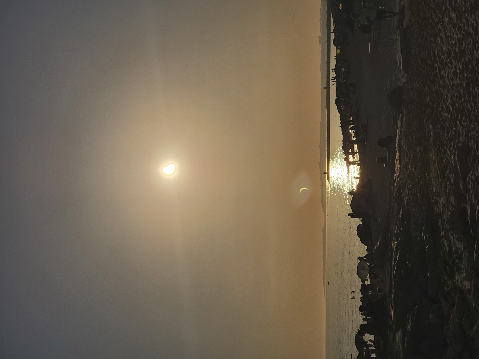 A partial solar eclipse is visible in the sky above Quarteira, Portugal, as the sun, partially obscured by the moon, casts a bright glare over the scene. The sun appears as a bright disc with a crescent shape cutout. The image captures a beach environment with numerous people scattered across the sand and in the shallow water. Some individuals are sitting under beach umbrellas, while others are standing or walking. The water reflects the sunlight, creating a shimmering effect. A breakwater or pier extends into the sea, with several figures visible on it. Silhouetted against the bright sky and water, the people and elements of the beach are rendered in dark tones. The sand is textured with ripples, suggesting a sandy shore. The overall lighting suggests either sunrise or sunset.