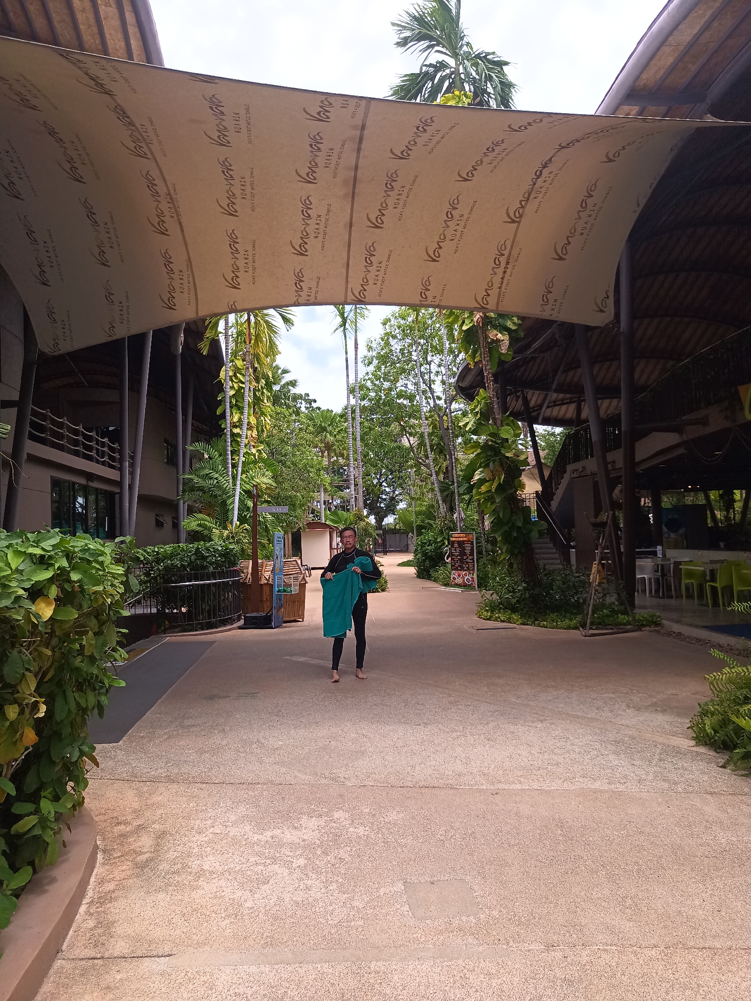 Here is a detailed description of the image:
**Scene:** The image captures a partially shaded outdoor pathway within what appears to be a resort or recreational area in Hua Hin City Municipality, Thailand. Overhead, there are large awnings bearing the words "Vana Nava Hua Hin" along with some text in a smaller font, suggesting this might be the Vana Nava Water Jungle.
**Environment:** Lush tropical plants and trees, including palm trees, are visible on both sides of the path. Buildings with architecture that blends with the tropical environment are also present. The ground is paved, and there are clear indications that this is a well-maintained and landscaped area.
**People:** A person, presumably a man, is standing in the center of the path, holding a green towel. He is wearing what looks like a black wetsuit or dark clothing and appears to be barefoot. He is looking directly at the camera.
**Objects:** Besides the awnings and vegetation, there are directional signs and other structures that are part of the resort. There are also glimpses of outdoor seating and bar areas, indicating recreational or dining facilities.
