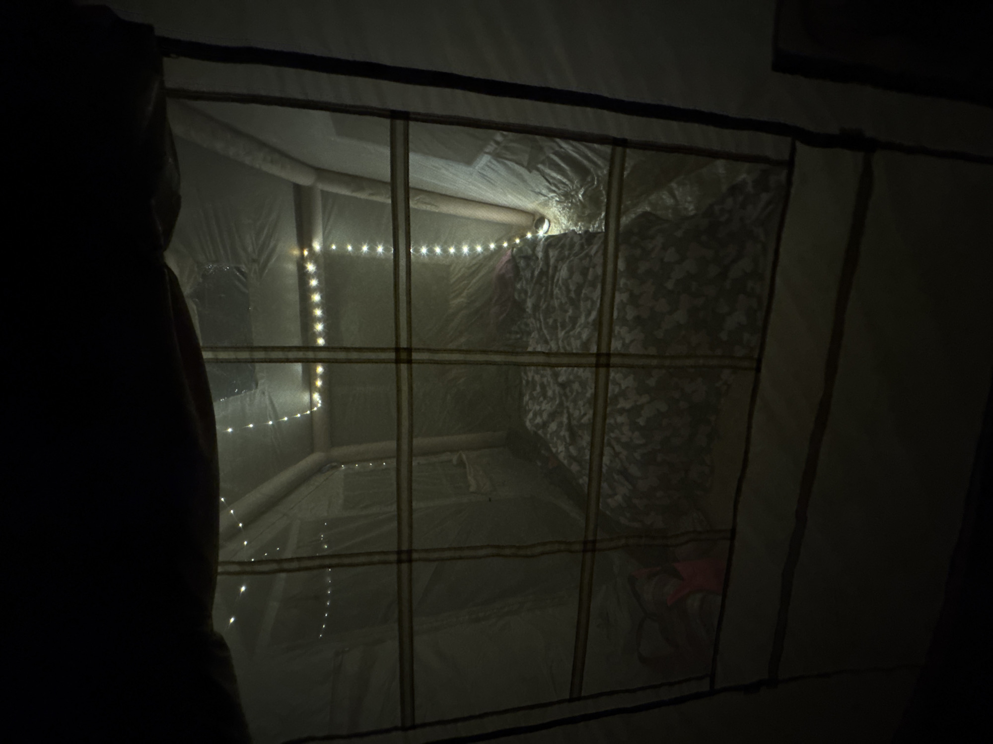 The image shows the interior of a tent, dimly lit by string lights. A bed covered with a floral patterned comforter is visible in the center. To the left of the bed, there are some pink and beige items, possibly bags. The tent walls are made of light-colored fabric. The scene appears to be set up for camping. The location is Zelenaya Roshcha, Russia.
