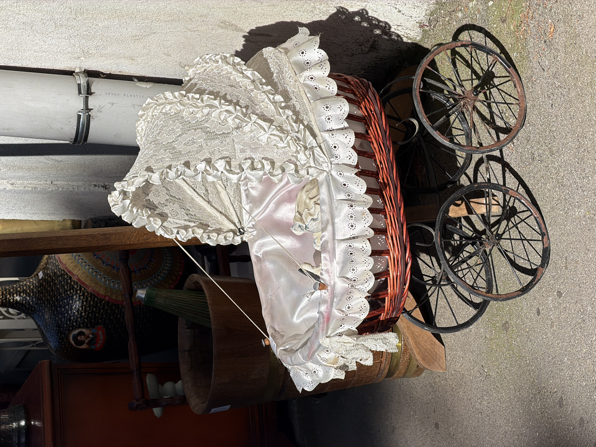 A high-angle shot depicts a vintage baby carriage with a white, lace-trimmed canopy and a wicker basket body. The carriage sits on a set of four iron wheels exhibiting rust. The carriage is located outdoors, likely on a paved surface in Tbilisi, Georgia. Behind the carriage, portions of what appears to be a cluttered display or storage area are visible, including wooden structures, a painted vessel, and a barrel. Overhead, a white plastic pipe with metal clamps is attached to the wall. The scene is lit by natural sunlight, casting shadows.