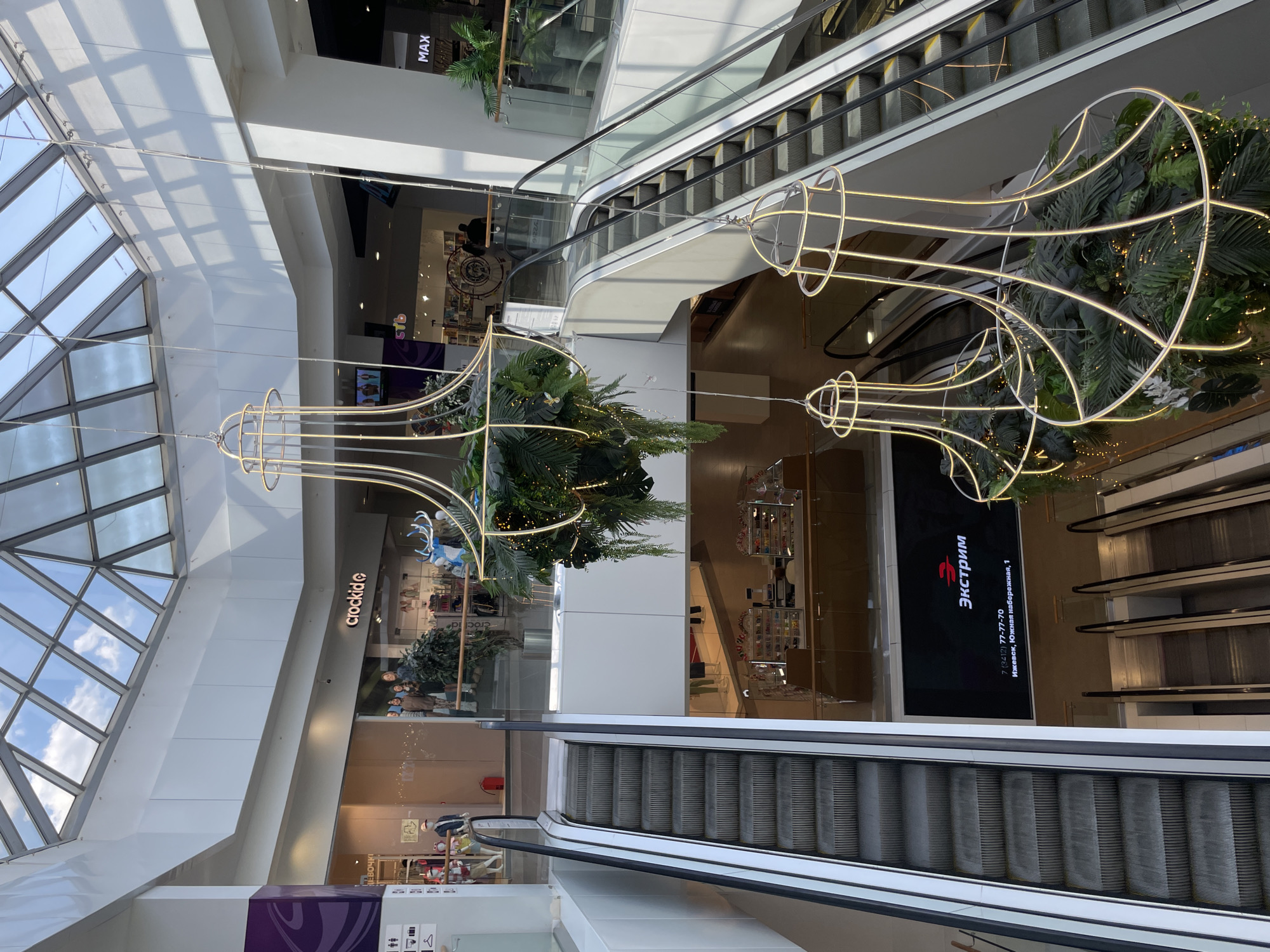 This interior shot shows an atrium in a shopping mall in Izhevsk, Russia. The view is from an upper level, looking down towards the lower levels. Daylight streams through a skylight. Escalators connect the floors. Decorative elements, resembling stylized trumpets with greenery inside, hang suspended in the open space. Storefronts are visible, including a “crockide” store. A large screen displays the word “Экстрим” and contact information for a business: phone number 7 (3412) 77-77-70 and the address, “Ижевск, Южная набережная, 1”, which translates to Izhevsk, Southern Embankment, 1. A "MAX" shop is visible in the upper left. People are visible through the storefronts.