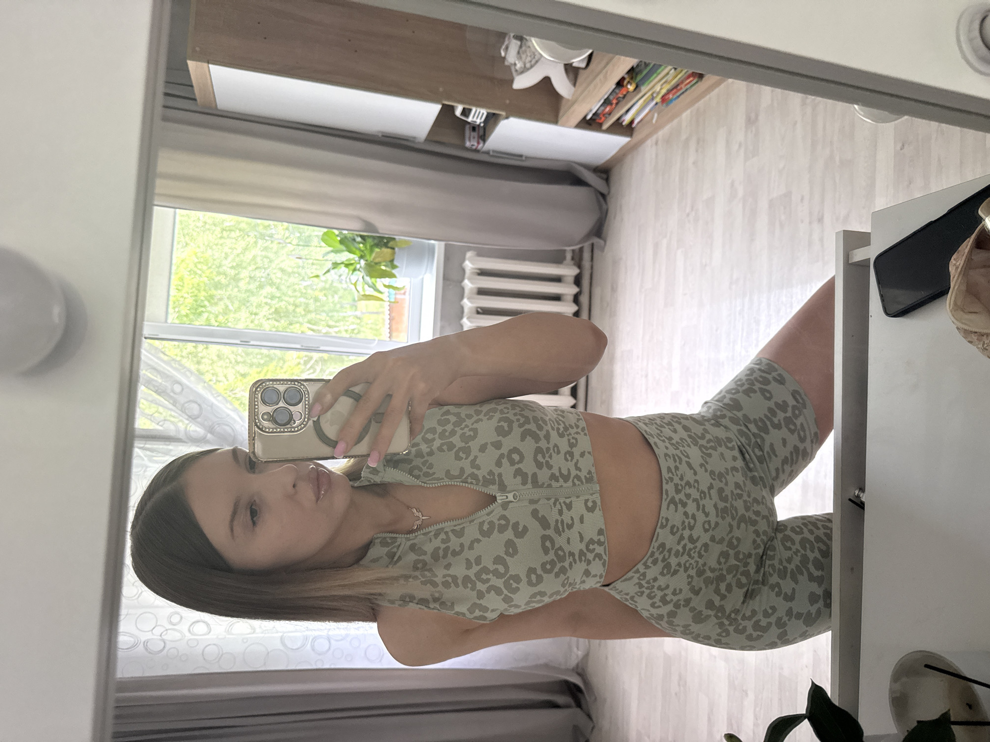 The media file shows a young woman taking a selfie in a mirror. She is wearing a light green leopard print athletic set, consisting of a cropped zip-up top and high-waisted biker shorts. She is holding a gold-colored phone with a decorative case. A delicate, thin necklace with a small charm is visible on her neck. Her hair is long and brown, styled straight with some lighter highlights.
The setting appears to be indoors, likely a bedroom or dressing area. In the background, reflected in the mirror, is a window with green foliage visible outside, suggesting daytime with pleasant weather. There is a radiator beneath the window, and a shelf unit with books and decorative items. The floor has a light wood laminate pattern. A white radiator is visible along one wall. To the right of the woman, a white surface, possibly a desk or dresser, is visible with a black smartphone and what appears to be a beige bra placed on it. The overall lighting is bright, indicating it is daytime. The woman's expression is neutral, focused on taking the photo. There is no visible text in the image. The location context is Октябрьское, Russia.