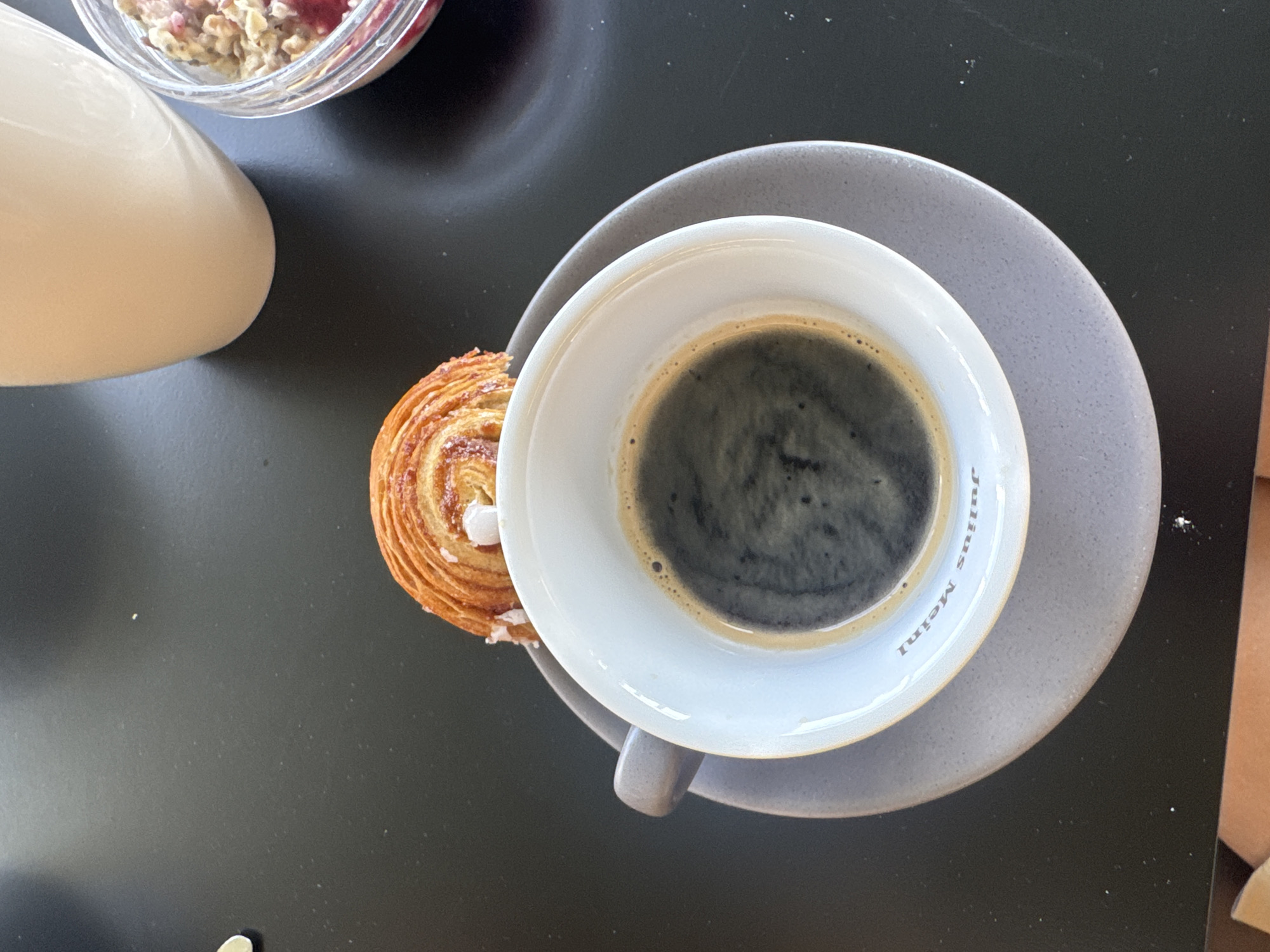 A close-up, overhead shot captures a breakfast scene on a dark, textured surface, likely a table. In the foreground, a white ceramic coffee cup sits on a matching grey saucer. The cup is filled with dark, frothy espresso, and the rim of the saucer has "Julius Meinl" subtly embossed on it. Resting atop the coffee cup's rim is a golden-brown, spiral-shaped pastry, appearing to be a kouign-amann or similar laminated dough, dusted with sugar and adorned with white icing or cream accents. To the right of the coffee cup, a clear glass bowl contains a breakfast medley of rolled oats, berries, and possibly nuts. Behind these items, the base of a cream-colored pitcher or mug is visible. The overall impression is a casual, appealing breakfast setting. The mention of Vienna, Austria, suggests this scene is likely in a café or restaurant within the city.