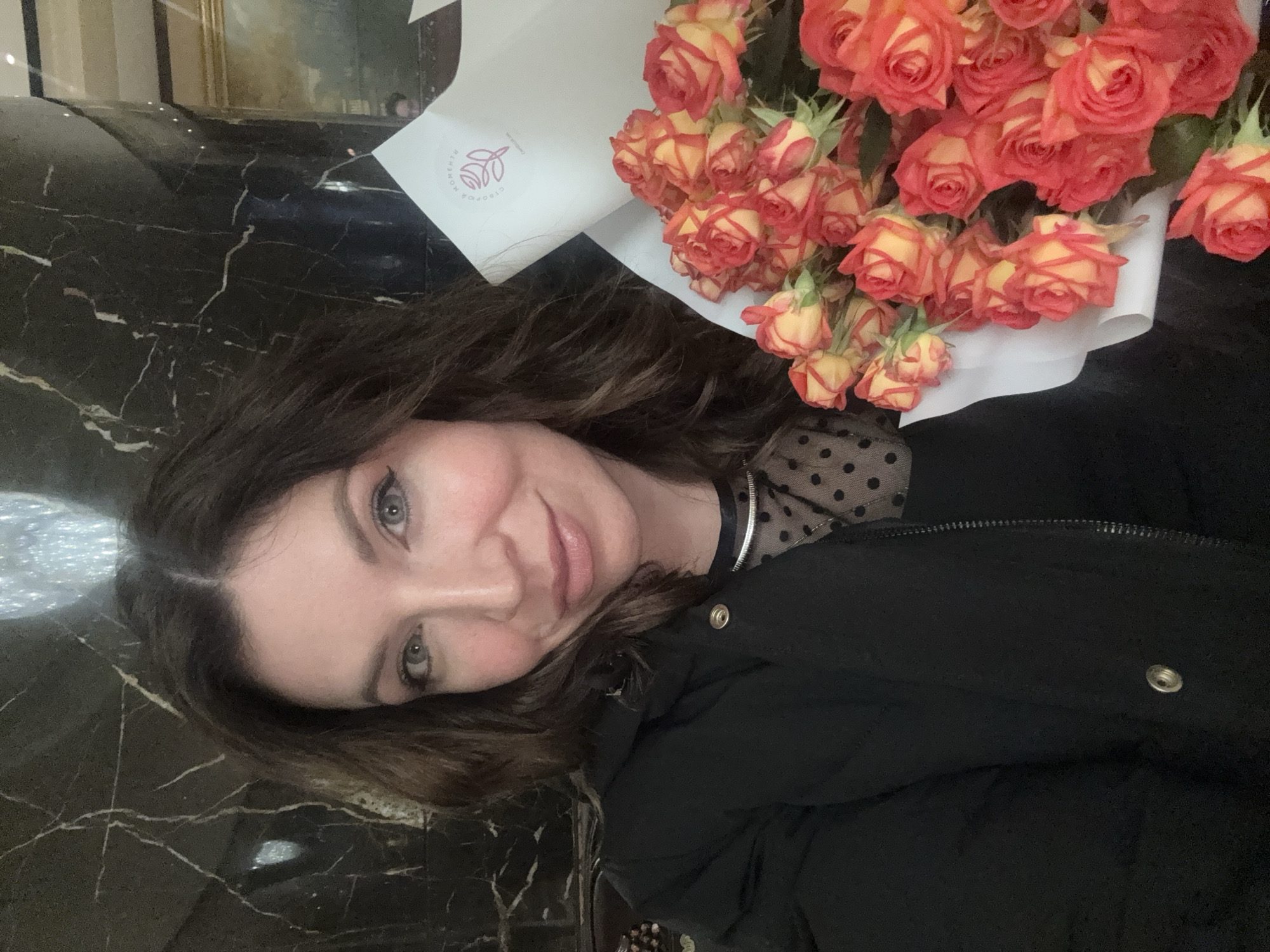 A woman poses with a bouquet of orange roses. She is wearing a black coat and a polka dot blouse with a silver necklace. The location appears to be indoors, with a black marble countertop in the foreground. The bouquet has a white paper wrap with a logo on it. The location is Unknown, Unknown.