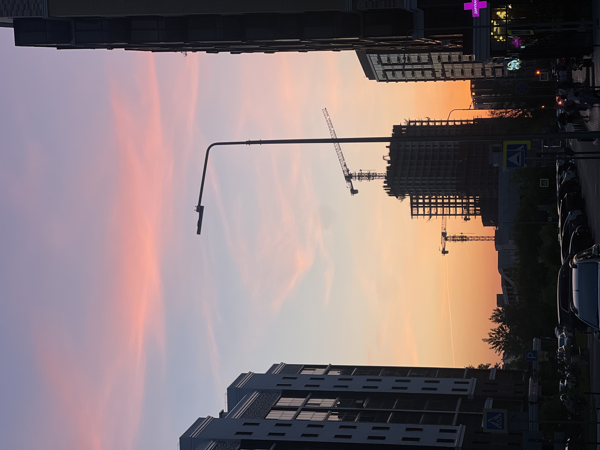 The photograph, taken in Kazan, Russia, showcases a cityscape at dusk. The sky is a gradient of lavender, pink, and orange. Two construction cranes stand silhouetted against the colorful sky above a construction site. Two modern buildings frame the top and bottom of the photograph. A street lamp is visible on the left side. Street signs, parked cars, and pedestrians are visible on the right.