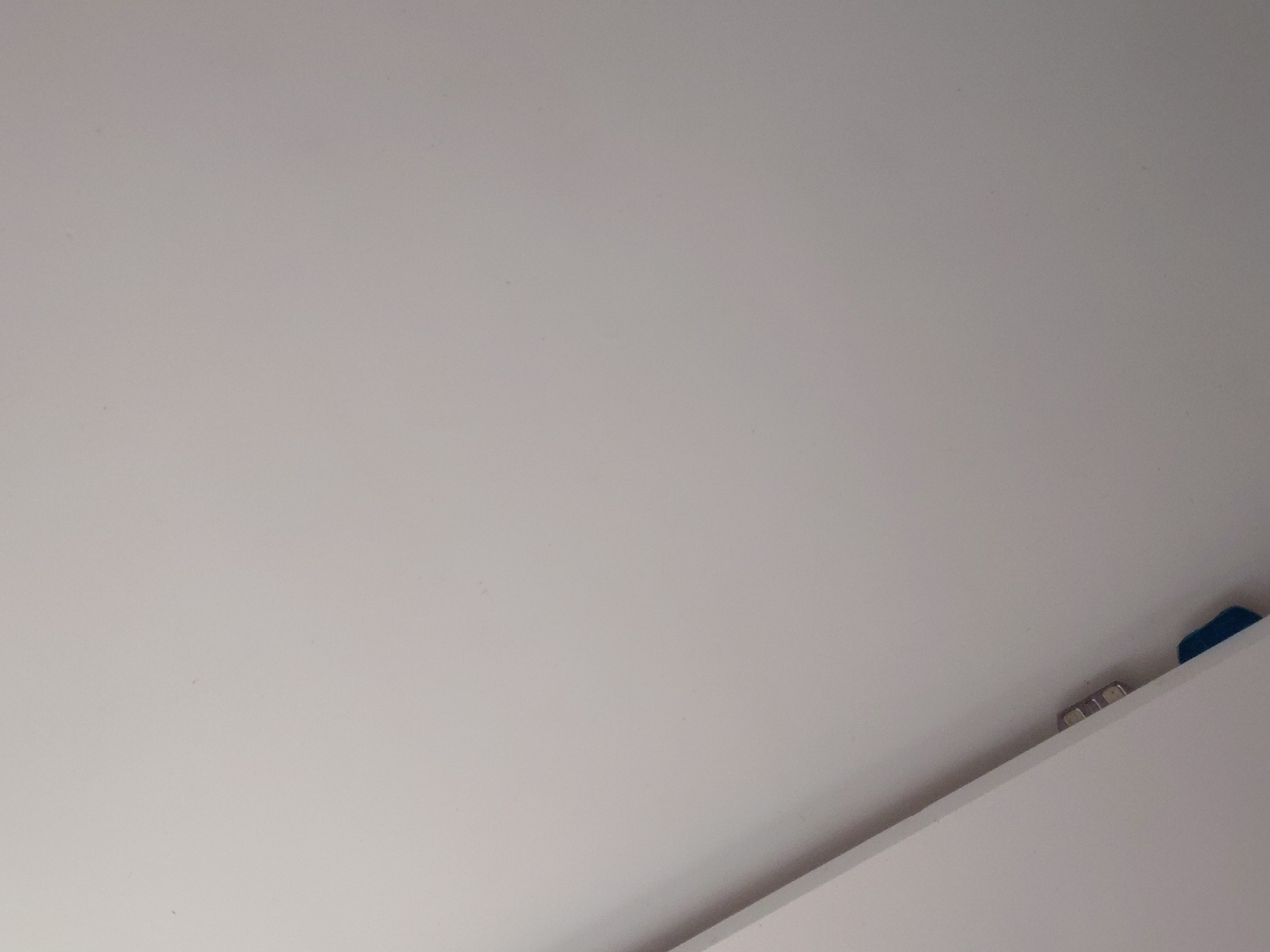 The image displays a close-up, upward-angled view of an interior architectural space, primarily dominated by a plain, light-colored surface, likely a wall or ceiling. This surface appears smooth with minimal texture, showing some very subtle specks or imperfections consistent with painted plaster or drywall.
In the bottom right corner, a sharp, diagonal line indicates the edge of another flat, white surface, possibly a shelf, cabinet, or part of another wall, creating a distinct boundary against the main background. Perched on this lower edge, partially visible, are a few small objects. There are two or three metallic, cylindrical items, possibly bolts, screws, or small magnetic fixtures, reflecting some light. To their right, a small, dark blue, rectangular object is visible, which could be a cap, a small container, or a piece of plastic.
No people or identifiable activities are present. The lighting is even and diffused, suggesting an indoor setting with either artificial light or indirect natural light, making it impossible to determine the time of day or external weather conditions. There are no discernible location cues within the image itself to suggest a specific geographical context. No text is visible anywhere in the frame. The overall impression is one of a minimalist and abstract composition, focusing on basic forms and a few small, incidental details.