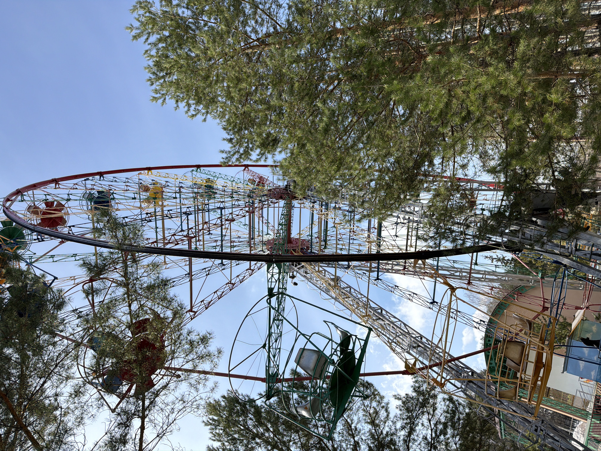 The image depicts a Ferris wheel in the city of Chaikovsky, Russia, taken from a low angle, looking up. The Ferris wheel is colorful with red, yellow, green, and blue elements. Some of the passenger cars appear to be old and possibly out of service. The Ferris wheel is partially obscured by pine trees. The sky is blue with some clouds.