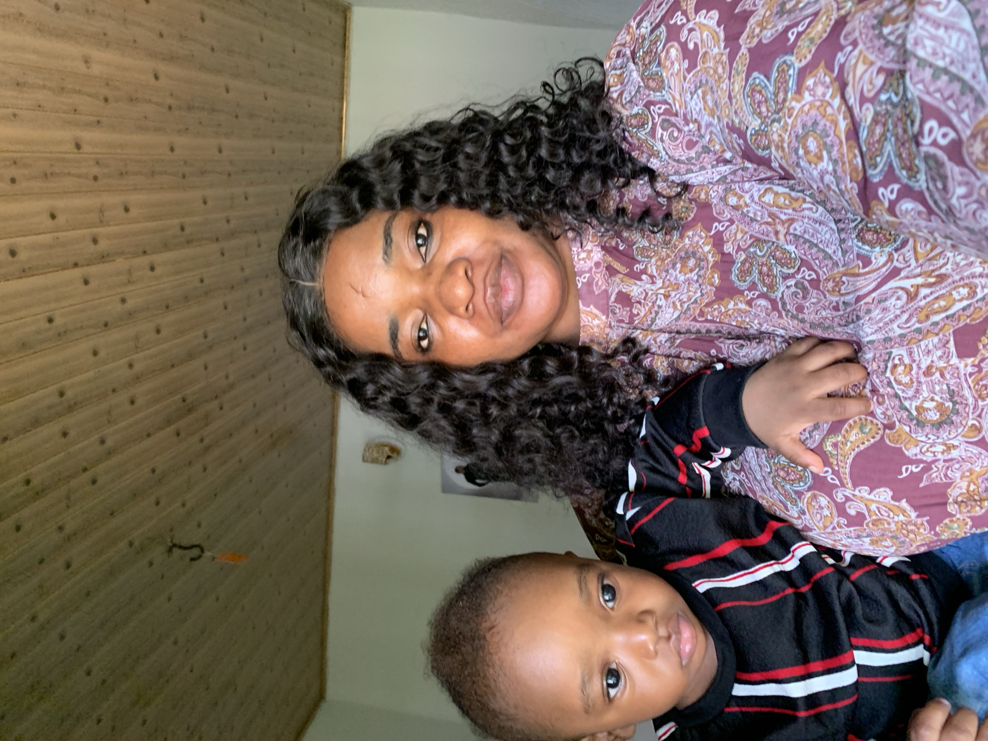 A selfie of a woman and a young child is taken indoors. The woman, who has dark, curly hair and is wearing a patterned maroon top, is positioned on the right side of the frame. The child, on the left, has dark hair and is wearing a black shirt with red and white stripes. The background shows a wooden plank ceiling and a plain wall. A hook with an orange object hangs from the ceiling. A framed picture is visible on the wall behind the child. The location is not specified in the image or accompanying text.