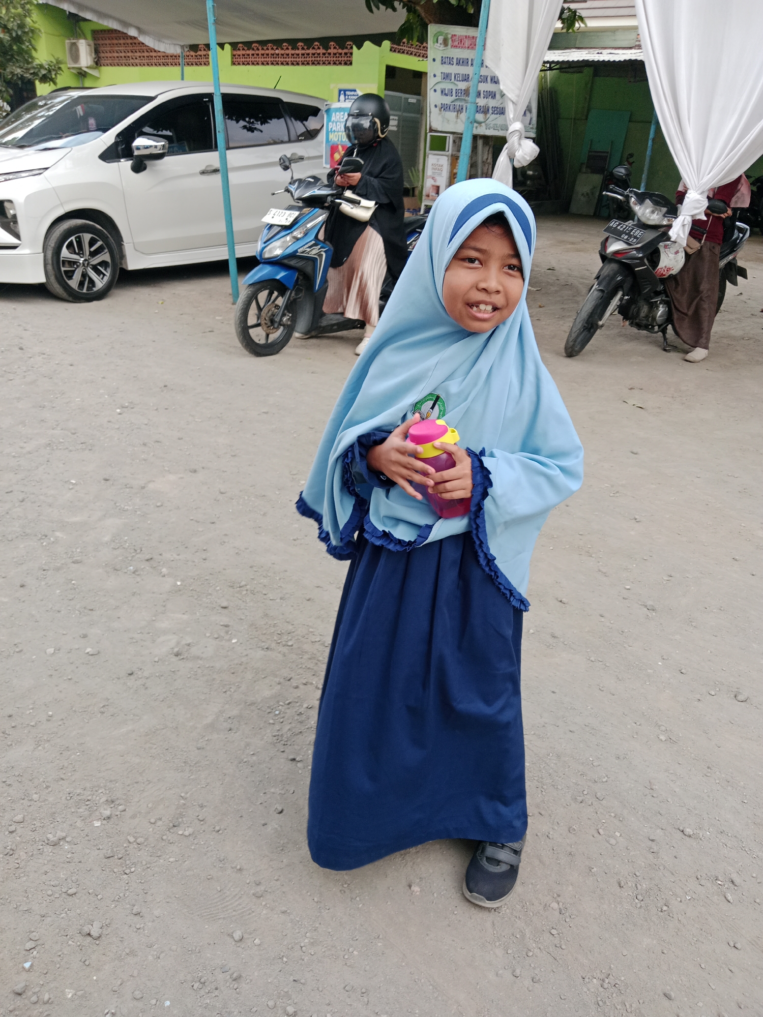 A young girl, approximately 7-9 years old, stands in the foreground, facing forward and smiling. She wears a light blue hijab with navy blue trim and a matching navy blue dress. She holds a pink and yellow water bottle in her right hand. Her left hand is visible near the bottle, and her feet are clad in dark grey velcro sneakers.
The background depicts an outdoor scene with a gravelly dirt ground. To the left, a white minivan is parked. Adjacent to it is a blue motorcycle with a person wearing a black helmet and a light-colored pleated skirt seated on it. Further to the right, a black motorcycle is parked, with a person wearing a brown skirt and a maroon top standing beside it. White tarps are visible in the background, suggesting a covered area. Signs with Indonesian text are also present on a green wall behind the motorcycles. The scene appears to be in a public area, possibly a parking lot or an event space, located in the city of Kediri, Indonesia.