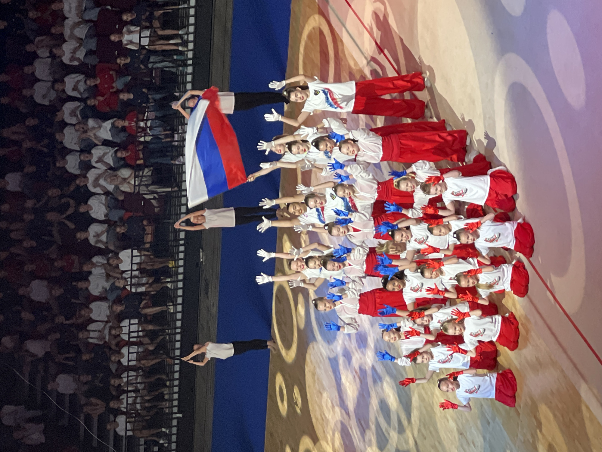 The image depicts a performance by a group of young gymnasts, likely during an event in Moscow, Russia. The gymnasts, dressed in red and white attire and wearing blue and red gloves, are positioned on a light-colored floor marked with circular patterns. They are arranged in multiple rows, with some kneeling and others standing, all with their arms raised. Two individuals in the background are holding a large Russian flag. Behind them, rows of spectators are visible in the stands, illuminated by stage lighting. The environment appears to be an indoor arena or stadium.