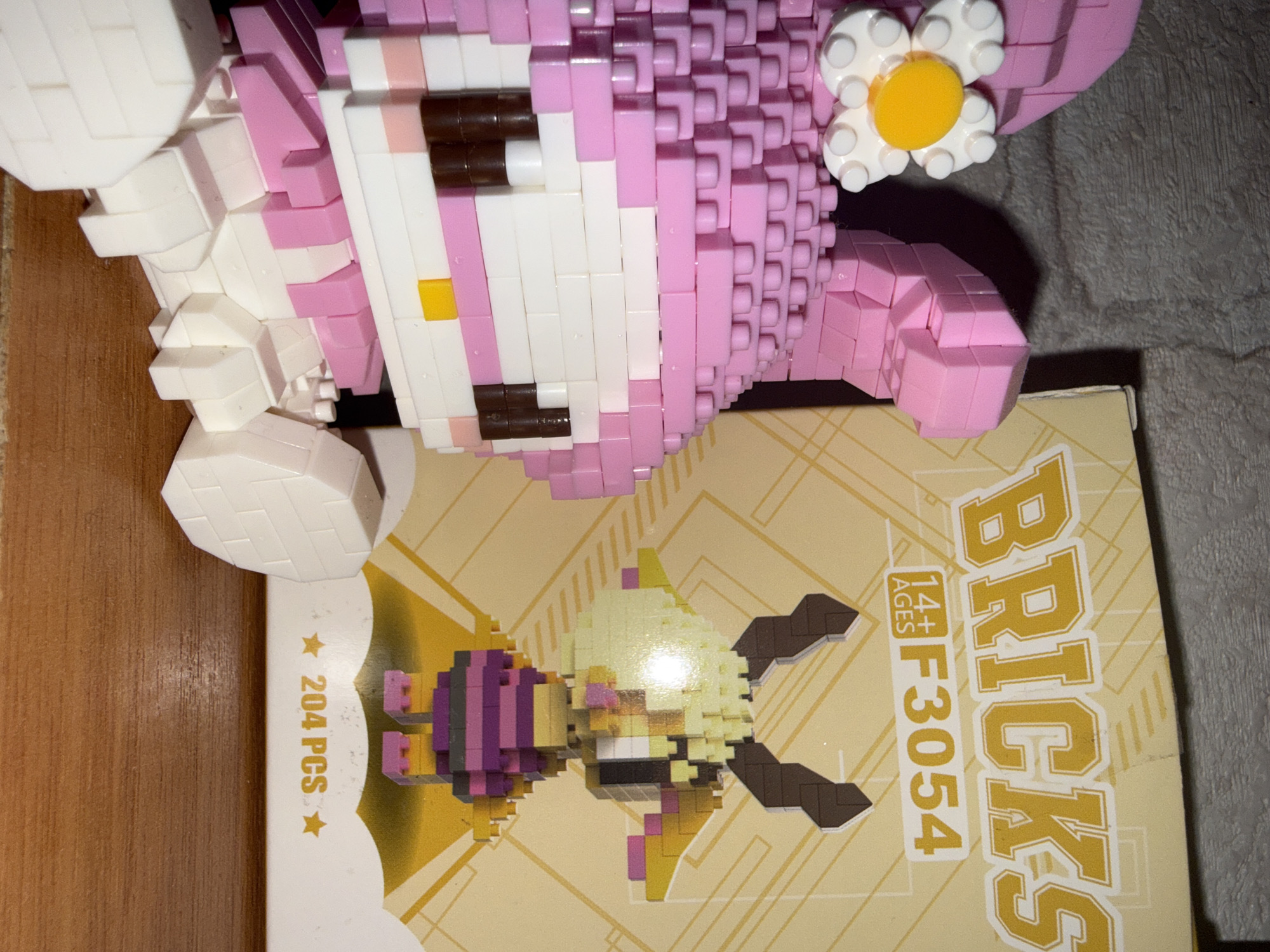 In Rayevskiy, Russia, a close-up shot focuses on a pink and white brick-built character resembling My Melody, with a white and yellow flower accessory on its head. To the right of the character is a portion of a yellow box labeled "BRICKS F3054" and "14+ AGES" along with an image of a brown and yellow brick-built character with horns. The box also indicates "204 PCS". The subjects are positioned on a wooden surface against a textured, dark background, possibly a wall.