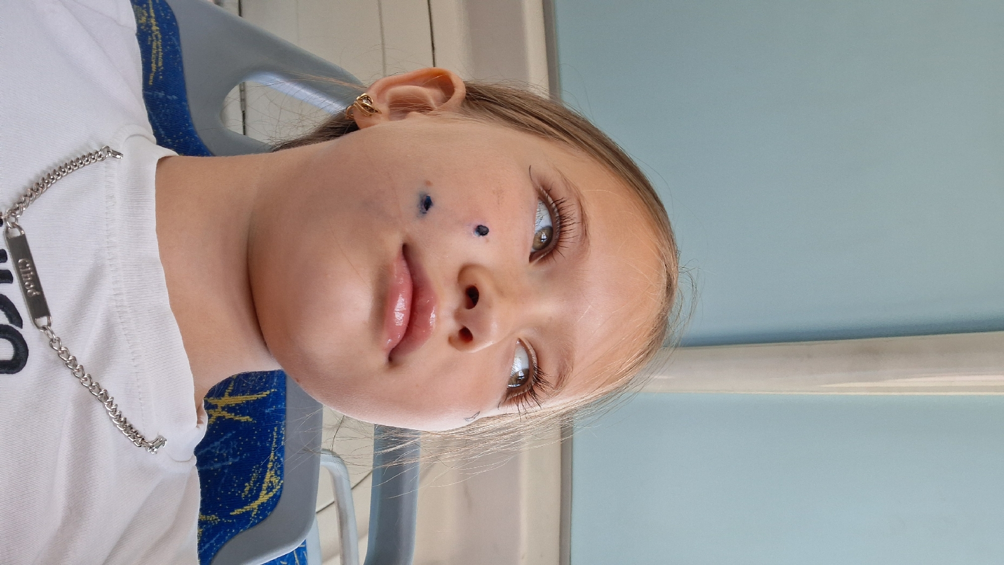A young Caucasian girl with blonde hair and green eyes is captured in a close-up, upward-angled shot. She is wearing a white t-shirt and a silver chain necklace. Her face is adorned with what appear to be blue smudges or temporary tattoos on her left cheek. She is also wearing a small gold hoop earring in her left ear. The background suggests she is seated, possibly on public transportation, with blue and gray seating visible behind her and a pale blue ceiling above. The scene appears to be indoors, with natural light illuminating the girl's face. The image does not contain any specific indicators of the city of Semey, Kazakhstan.