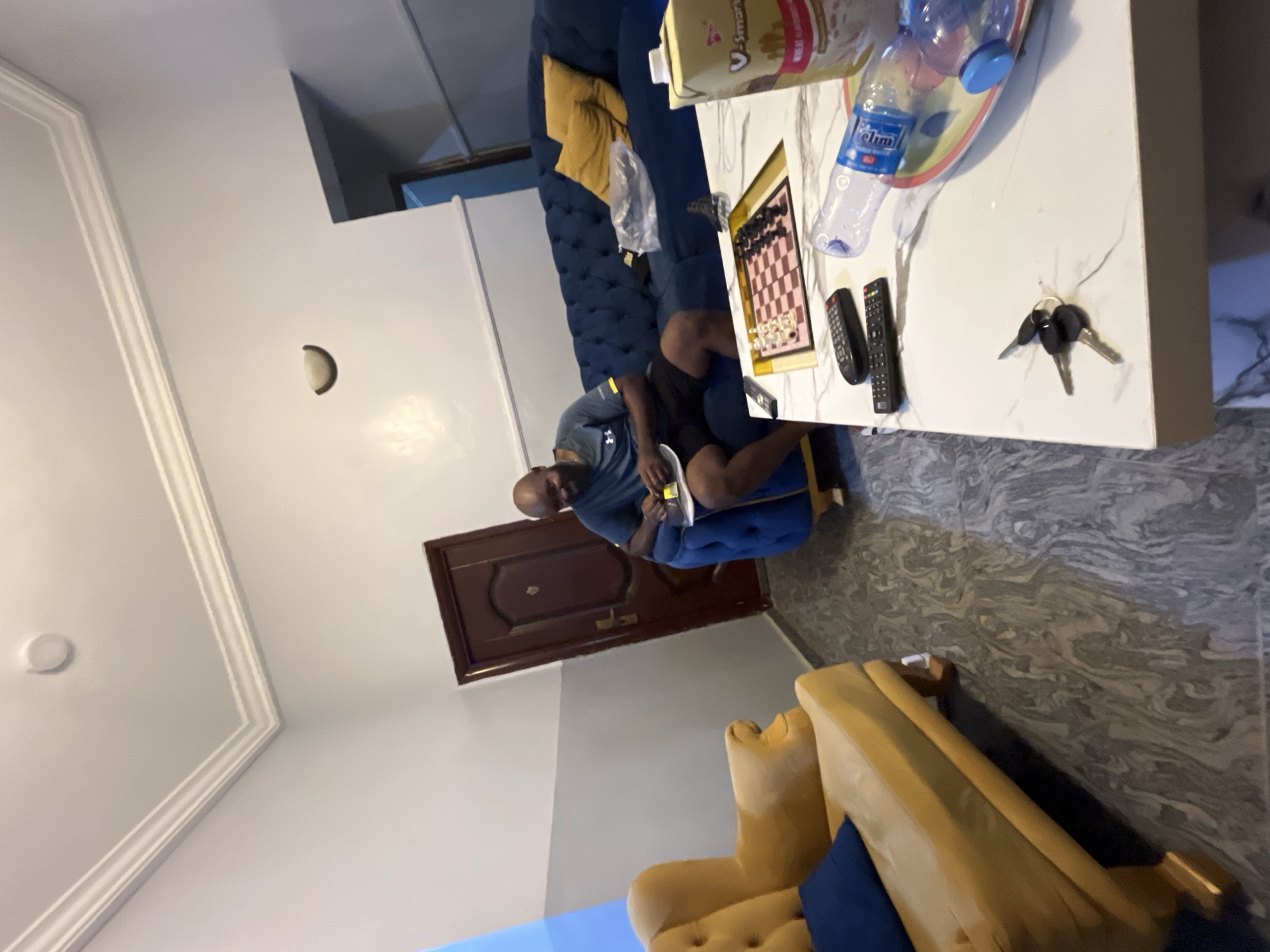 The image captures an indoor scene, likely a living room, in Dushepe, Nigeria. The primary subject is a man, bald and wearing a blue t-shirt and dark shorts, seated on a blue upholstered sofa. He is holding a small, white-covered item, possibly a phone or notepad, and appears to be engrossed in it.
The room is furnished with a tufted blue sofa, where the man is seated, and a lighter-colored, possibly yellow or beige, armchair is partially visible in the foreground. A white coffee table with a marbled pattern sits in front of the blue sofa. On the table, several items are arranged: a chessboard, a plastic water bottle with the brand "Elim" visible, a cereal box labeled "V-Smart Wheat Supplement," two black remote controls, and a set of keys.
The walls are painted white, featuring decorative ceiling molding. A brown wooden door is visible in the background, contrasting with the lighter walls. The floor is covered with gray tiles displaying a swirled, marbled pattern. The lighting suggests daytime indoors, possibly with artificial light sources as no windows are clearly visible. The man's demeanor seems calm and relaxed. A part of a white appliance, possibly a refrigerator, can be seen in the background on the left.
