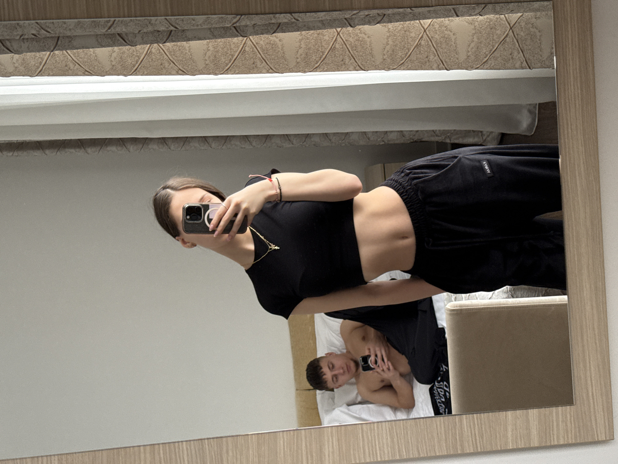 The image depicts an indoor scene in Vozdovac Urban Municipality, Serbia, captured as a mirror selfie. A young adult female stands in the foreground reflection, holding a dark-colored smartphone to take the photo, obscuring her upper face. She is dressed in a black short-sleeved crop top, revealing her midriff, and black sweatpants with a visible white 'AMRAX' label on the left thigh. She wears a gold chain necklace with a small cross pendant, a ring on her left ring finger, and multiple thin bracelets on her left wrist. In the background reflection, a young adult male is seated on a bed with white bedding, propped against a light-colored upholstered headboard. He is shirtless, wearing dark trousers, and is focused on a dark-colored smartphone he holds with both hands. A light-colored upholstered chair is partially visible in the lower-left reflection. To the right, a window is covered by an inner sheer white curtain and an outer beige patterned curtain with a diamond-like geometric design. The walls of the room are a plain light color.