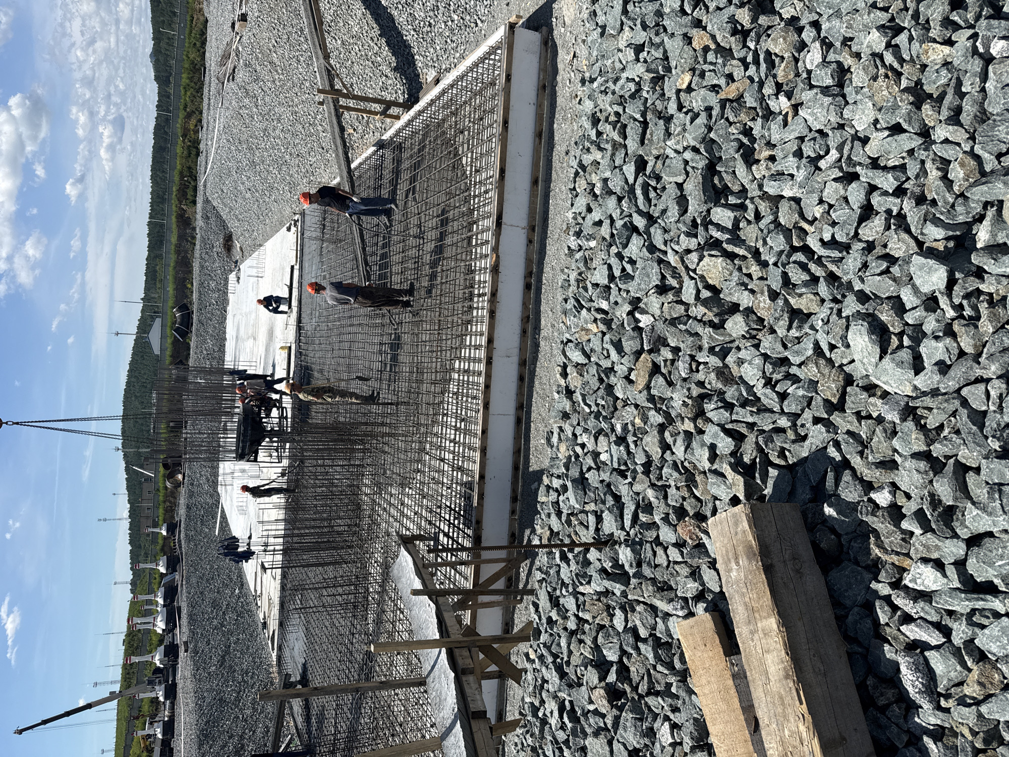 In the city of Leninsk, Russia, a construction site is depicted under a clear sky with scattered clouds. The foreground is dominated by a large pile of grey gravel. In the midground, a concrete foundation is being prepared. Reinforcement bars form a dense grid over the foundation, with workers in hard hats actively engaged in construction tasks. A crane is visible in the background, lifting a concrete bucket. Several other cranes and white structures are also present further in the distance, alongside a line of trees. The overall scene suggests ongoing infrastructure development.