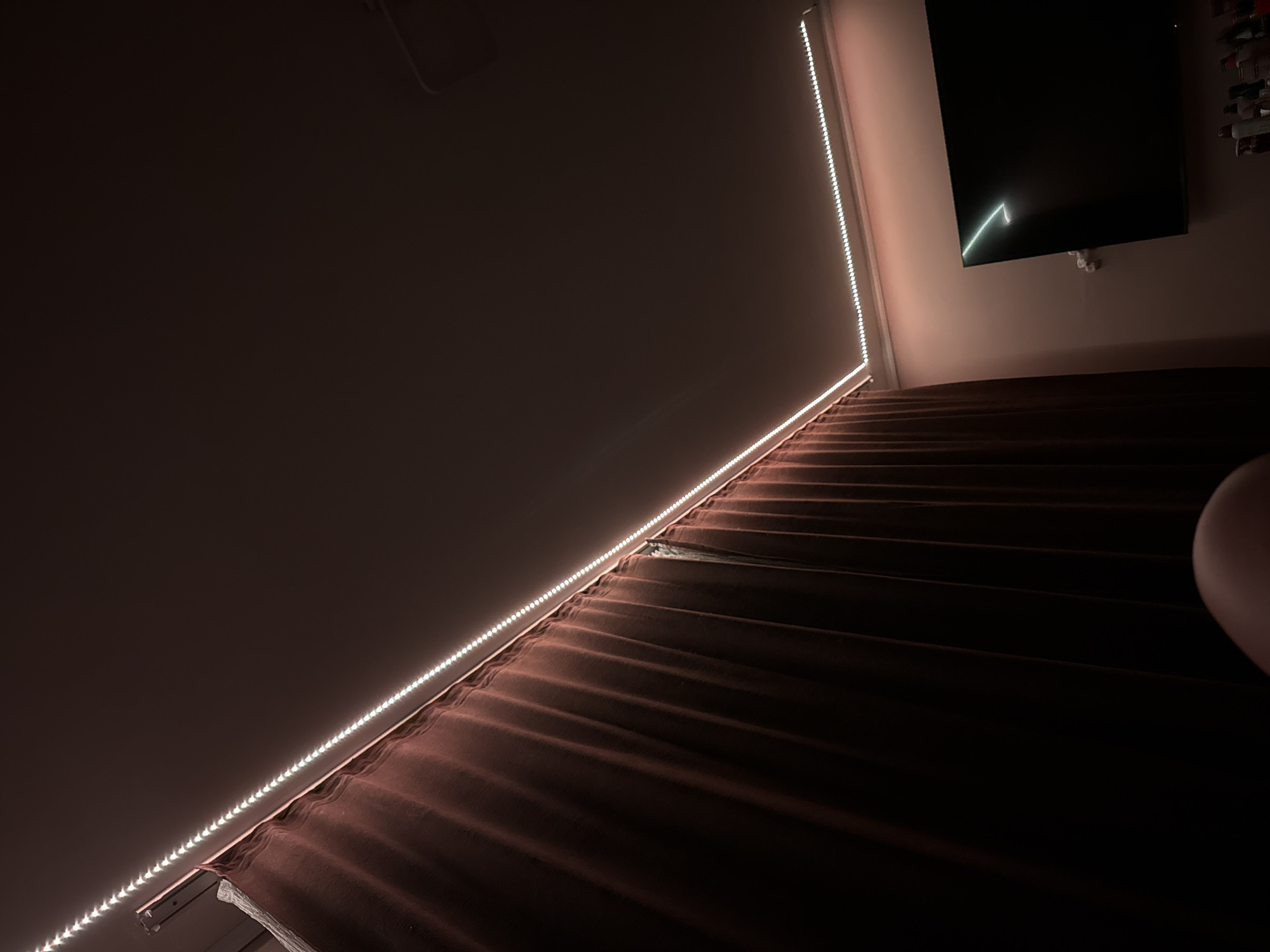 The image presents an indoor scene, likely a room at night or in low light, dominated by artificial illumination. The composition is angled, creating a strong diagonal line across the frame.
The central subjects are bright, white LED light strips that run along a light-colored wall. One strip extends diagonally from the lower left to the center, and another continues vertically upwards along the right side of the wall. These lights cast a soft glow on the wall, which appears to be a light off-white or cream color, and illuminate the textures of the room.
Below the diagonal LED strip, a large expanse of dark, possibly brown or burgundy, pleated fabric is visible. This fabric, likely curtains or bedding, displays distinct folds and shadows, creating a sense of depth and texture. In the upper right corner, a dark, rectangular television screen is mounted on the wall. A thin, angular, light-colored line is faintly visible on its surface, which could be a reflection or part of a screen display. Mounting brackets are visible beneath the television. To the far right, partially obscured by shadow, is a shelf or rack holding several small, rectangular objects. A rounded, light-colored object is also partially visible at the extreme right edge of the frame.
There is no specific activity taking place; the image captures a static interior view. The time of day appears to be evening or night due to the reliance on artificial lighting and the dark surroundings. The lighting creates a serene, almost contemplative atmosphere. No text is visible within the image. While the location context is given as Rogosh, Bulgaria, no specific visual cues within the image confirm this geographical detail.