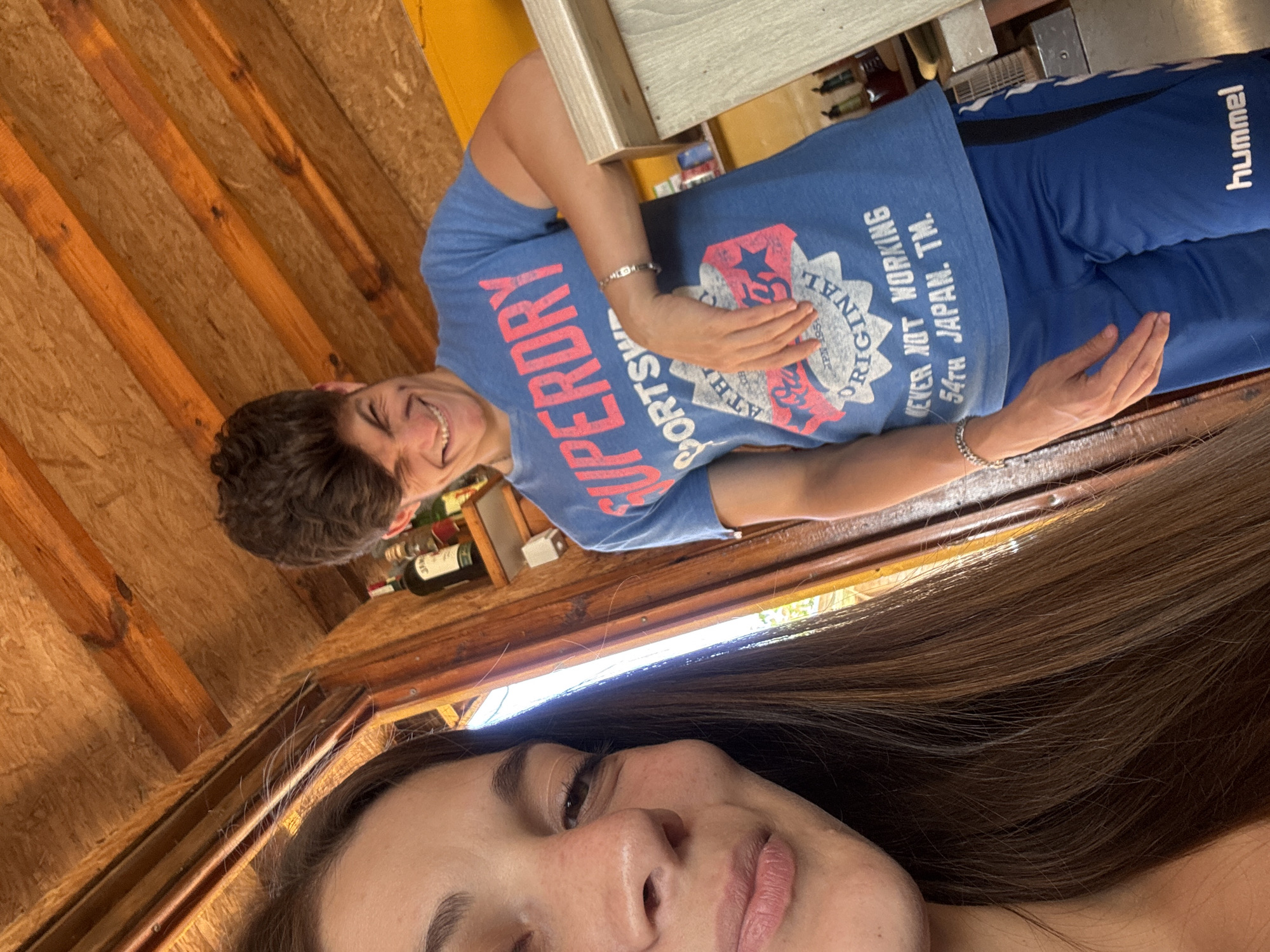 The image shows a young woman and a young man standing inside a wooden structure. The woman is in the foreground, with her face partially visible on the left side of the frame. She has long, dark brown hair and is looking towards the camera with a slight smile. The man is in the background, on the right side of the frame. He has short, dark, curly hair and is wearing a blue t-shirt with text and graphics, and blue athletic shorts with a white logo. He is leaning on a counter and smiling broadly at the camera. The background shows wooden walls and ceiling, and shelves with bottles. The setting appears to be a casual indoor space, possibly a bar or a shop. The overall lighting suggests it is daytime. The image is taken from a low angle, with the camera pointed upwards.