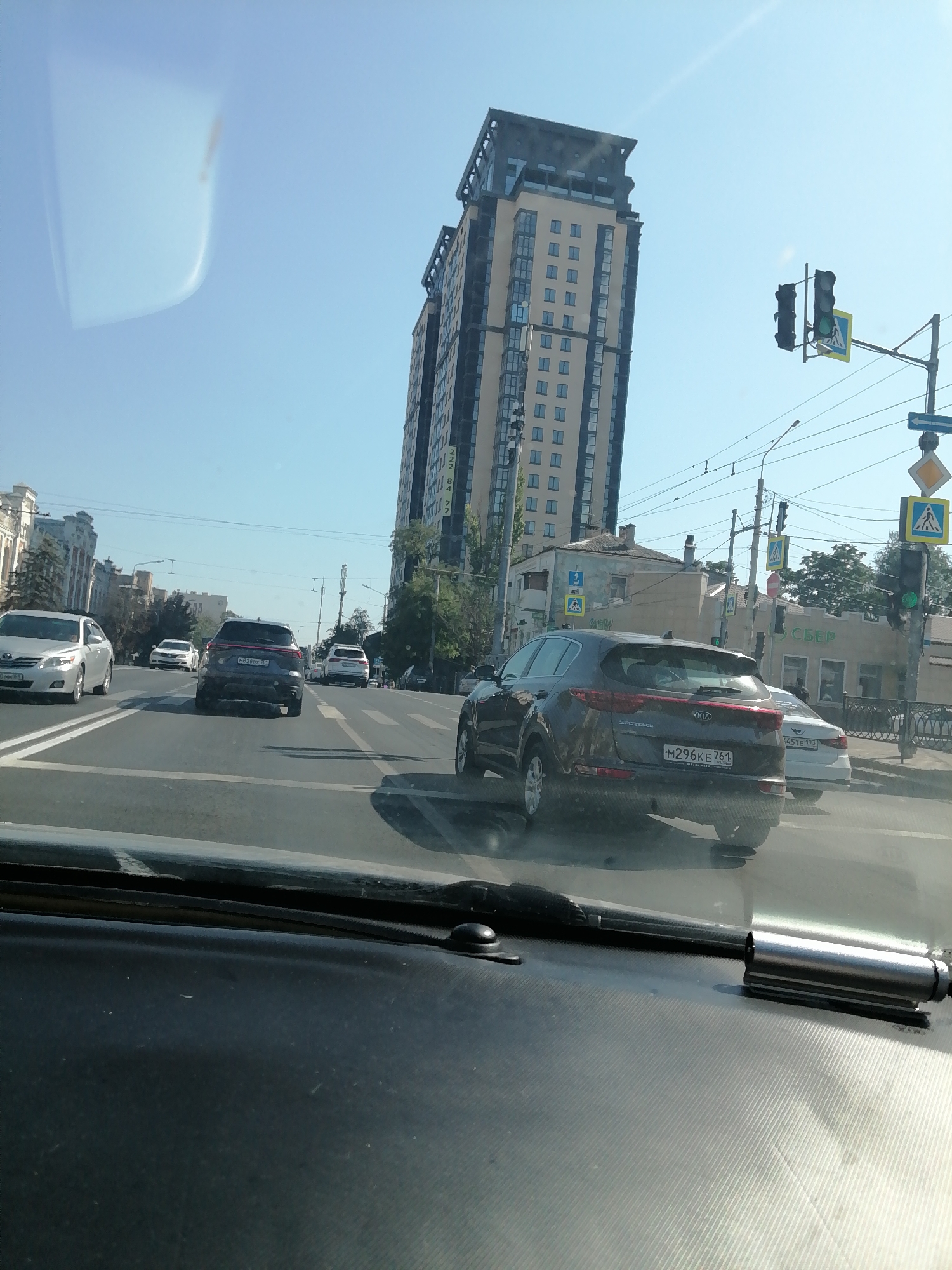 The image captures a view from inside a vehicle, looking out at a city street in Rostov-on-Don, Russia. The scene unfolds under a clear blue sky, with a tall, modern residential building dominating the background on the right. This building features a light-colored facade with numerous windows. To the left of this building, several other structures of varying architectural styles are visible, including older, more ornate buildings and what appears to be a commercial building with a prominent green sign.
The street is active with vehicular traffic. Several cars are visible, including a dark SUV in the foreground with the license plate "M296KE 761". Other cars include a silver sedan and a white SUV. Traffic lights are present, with one clearly showing a green light. Pedestrian crossings are marked on the road, and pedestrian crossing signs are also visible. Overhead power lines crisscross the sky, indicating the presence of infrastructure typical of an urban environment. The foreground of the image shows the interior of the vehicle, including the dashboard and windshield wipers. The overall impression is of a typical day on a busy city street.