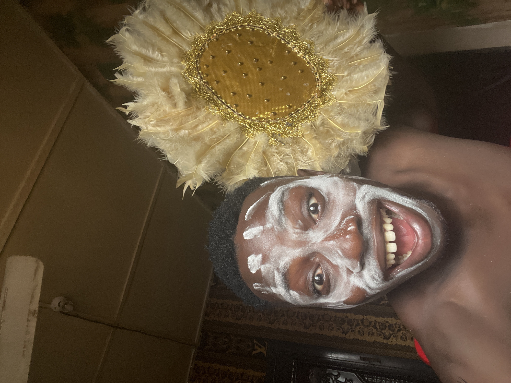 The media file features a person, likely male, with a wide, joyful smile, revealing their teeth. Their dark skin is adorned with white ceremonial paint applied in distinct patterns, including lines on the forehead, around the eyes, and across the cheeks and nose. They have short, dark, curly hair.
The person wears an elaborate, circular, fan-like headpiece composed of numerous light beige or off-white feathers radiating from a central, gilded disc. This disc is textured, possibly made of gold fabric, and decorated with small golden studs or beads, further framed by ornate gold lace or trim. A hint of a red garment or accessory is visible near their shoulder, suggesting traditional attire.
The image is oriented sideways. The background, when viewed upright, includes dark brown wall paneling on one side and a dark, patterned rug or carpet on the other, displaying what appears to be an "LV" (Louis Vuitton) monogram. The setting is indoors and dimly lit.
The facial markings, elaborate headpiece, and joyful expression suggest the person is participating in a cultural ceremony, traditional performance, or festive event in Nkpor, Nigeria.