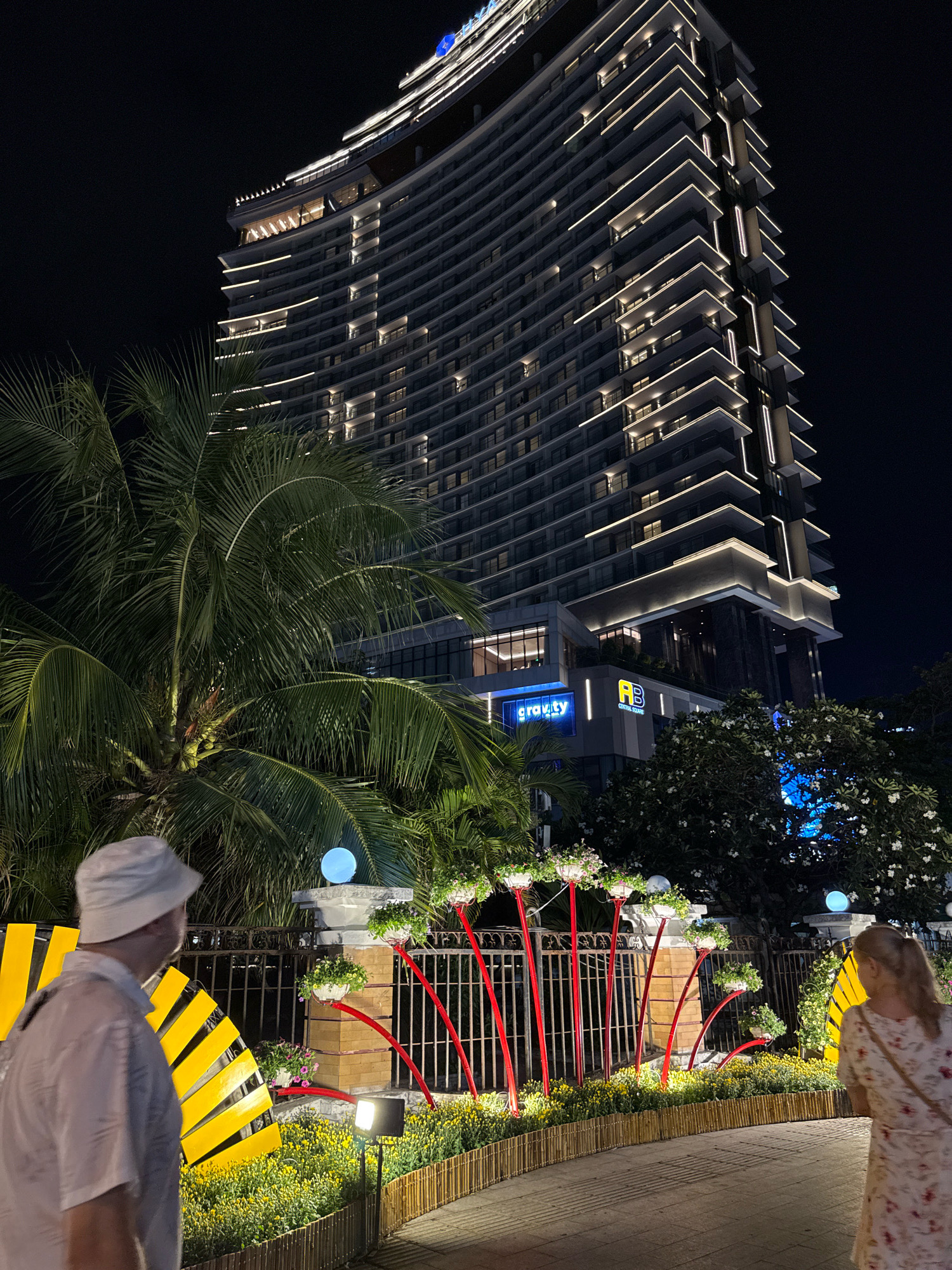 This is a night-time scene in Nha Trang, Vietnam, featuring the AB Central Square building and its surrounding area. The tall, modern building is illuminated with horizontal white lights along its many floors, creating a striking architectural display against the dark sky. In the foreground, a paved walkway is lined with a low bamboo fence and filled with yellow flowers. To the left, a person wearing a white hat and a light-colored shirt is seen from the back, facing away from the camera. On the right, another person, a woman wearing a floral dress, is also viewed from the back, walking away. Between the people and the building, a decorative fence with red curved elements supports planters filled with greenery. Palm trees are visible to the left of the building, and a bright blue spherical light fixture sits atop a pillar behind the fence. A neon sign reading "gravity" is visible on the building. The overall impression is a well-lit urban environment at night with a focus on modern architecture and decorative landscaping.