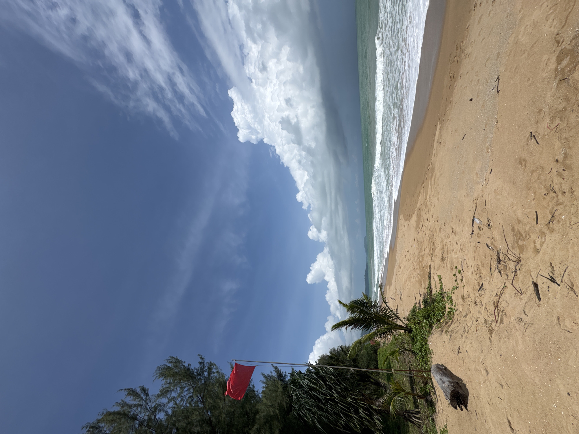 A wide-angle, low-angle shot captures a tropical beach scene in Ban Khuk Khak, Thailand. A bright red flag flies from a pole on the left side of the frame, near a cluster of dark green trees and palm fronds. The sand in the foreground is light brown, with some scattered driftwood and debris. Gentle waves roll onto the shore, with the ocean stretching towards the horizon. The sky above is a vibrant blue, dotted with white, fluffy clouds and wispy cirrus formations. In the distance, the coastline is visible, with more trees and landmasses. The overall impression is one of a serene, natural environment, though the red flag suggests a warning or caution.
