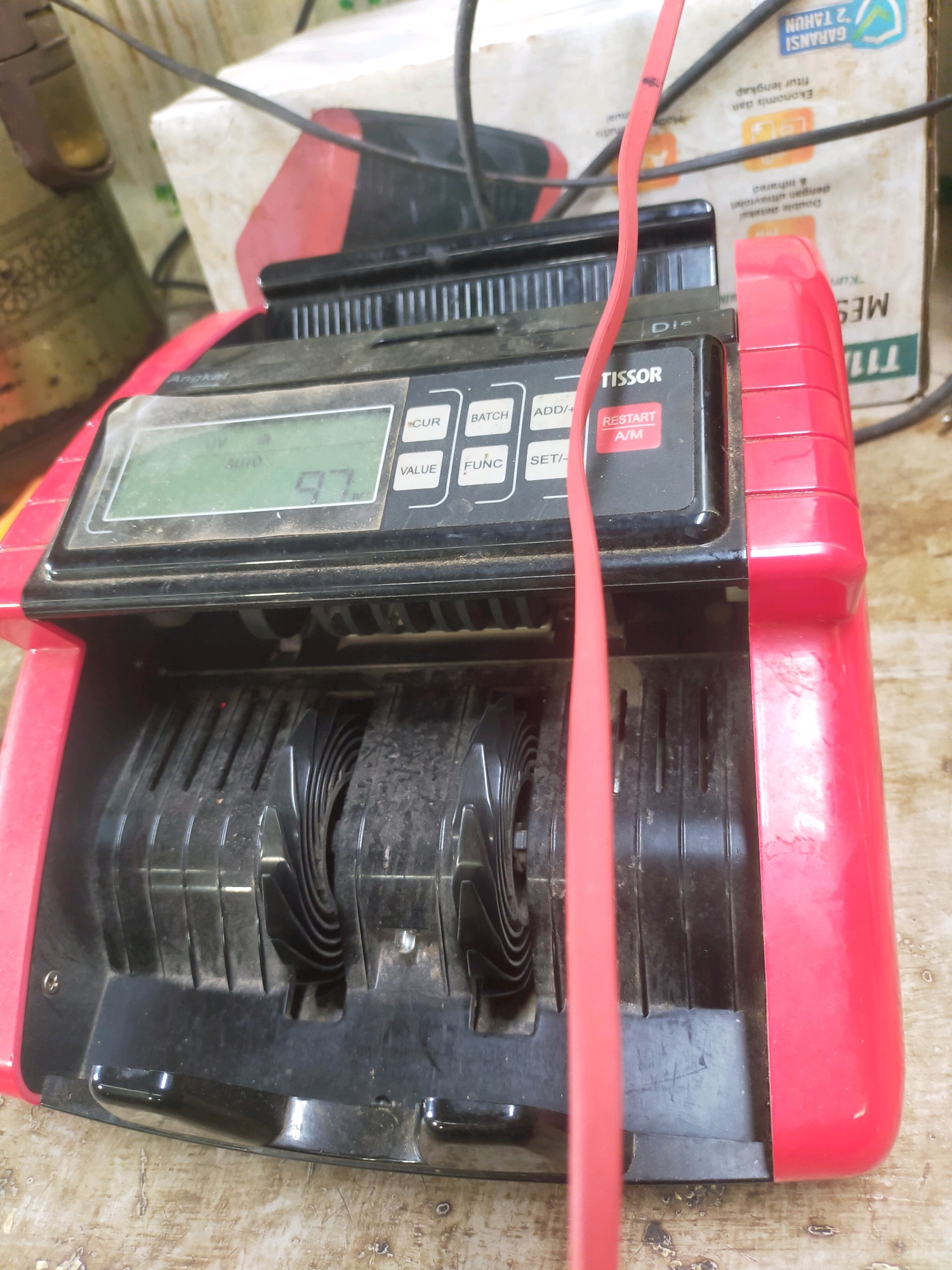 A red and black electronic currency counting machine, identified as a "Tissor," is the primary subject in the foreground of the image. The machine displays the number "97" on its LCD screen, indicating a count. Several buttons with labels such as "CUR," "BATCH," "ADD/+", "VALUE," "FUNC," "SET/-," and "RESTART A/M" are visible on the control panel. A bright red cable is draped across the top of the machine and extends downwards. The machine rests on a weathered wooden surface with visible scratches and peeling paint. In the background, a cardboard box with Indonesian text, including "GARANSI 2 TAHUN" (2-year warranty) and "MES T11", is partially visible. The overall environment appears to be indoors, possibly a workshop or a retail space, with hints of other objects and a slightly dusty atmosphere. The image location is specified as Mojodanu, Indonesia.