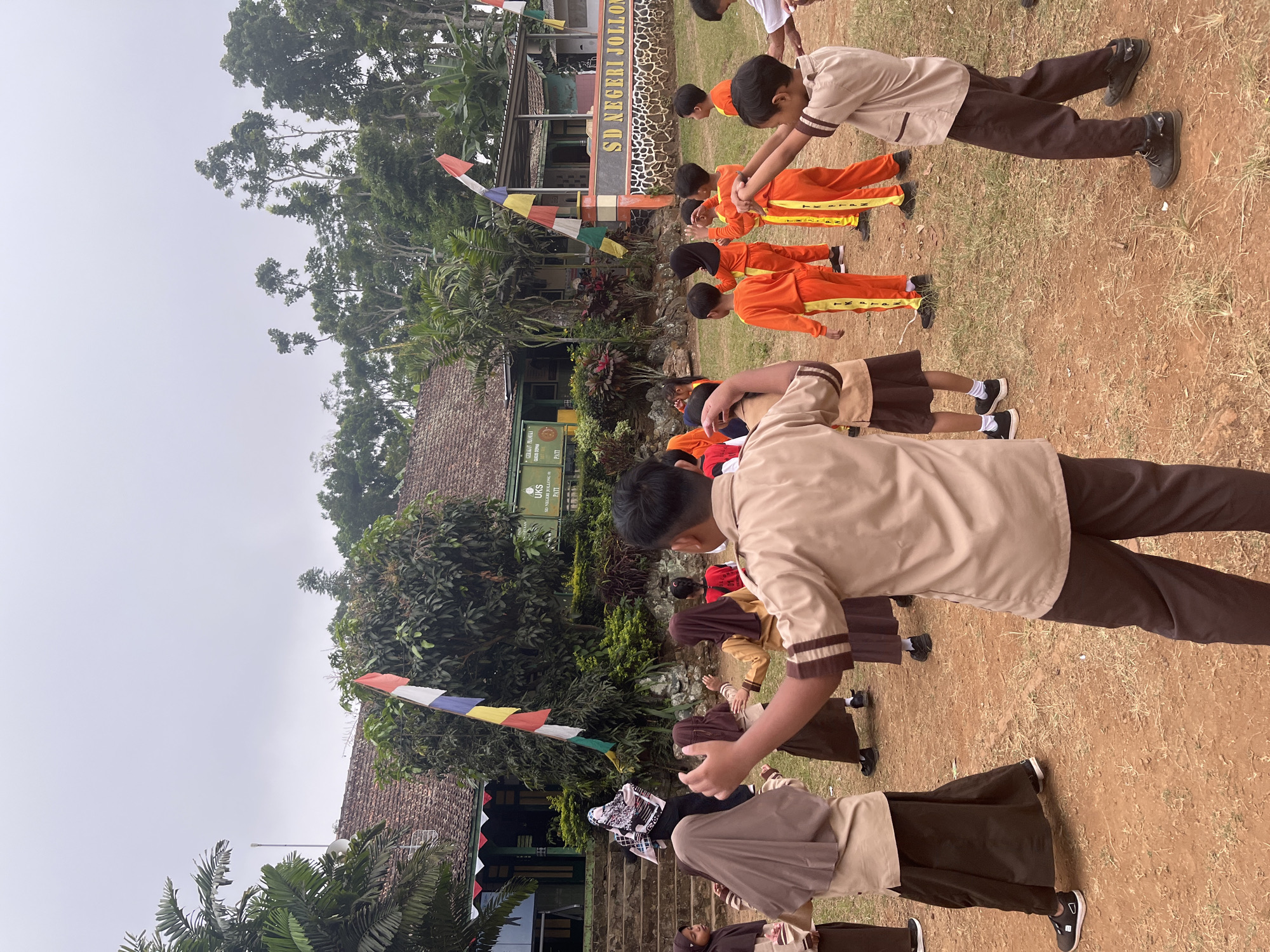 In Gembong, Indonesia, a group of elementary school children in uniform are engaged in physical activity outdoors. The scene depicts a schoolyard with a building in the background, featuring traditional tiled roofs and banners, including the Indonesian flag. Several children, both boys and girls, are dressed in beige and brown uniforms, while others wear bright orange tracksuits. They appear to be following instructions, possibly from an unseen teacher or instructor, with some children performing coordinated movements. The ground is covered in dirt and scattered dry grass, indicating an outdoor setting. The overall atmosphere suggests a typical school day activity, possibly a physical education class or a morning exercise routine, under an overcast sky.
