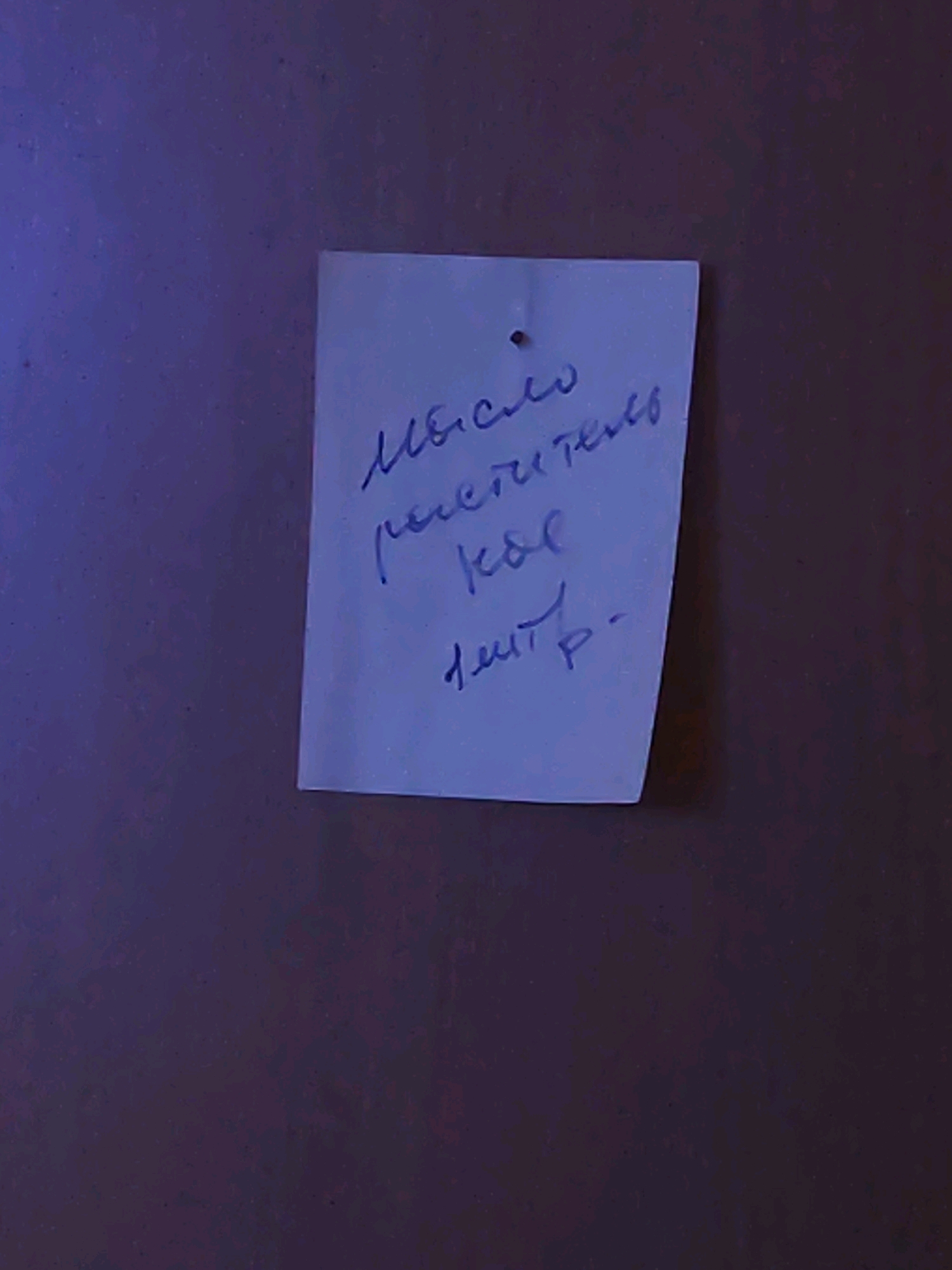 A close-up, low-angle shot captures a white, slightly creased note with handwritten text attached to a dark, textured surface, possibly a wall or door. The text, written in blue ink in a cursive, handwritten script, appears to be in Russian. The note is pinned to the surface by a single tack. The lighting in the image is dim and casts a purplish hue, suggesting it was taken indoors or at night. The background is out of focus and predominantly dark with some subtle variations in tone. The city of Balqaş, Kazakhstan, is not visible in this image, and no information about its location is present.