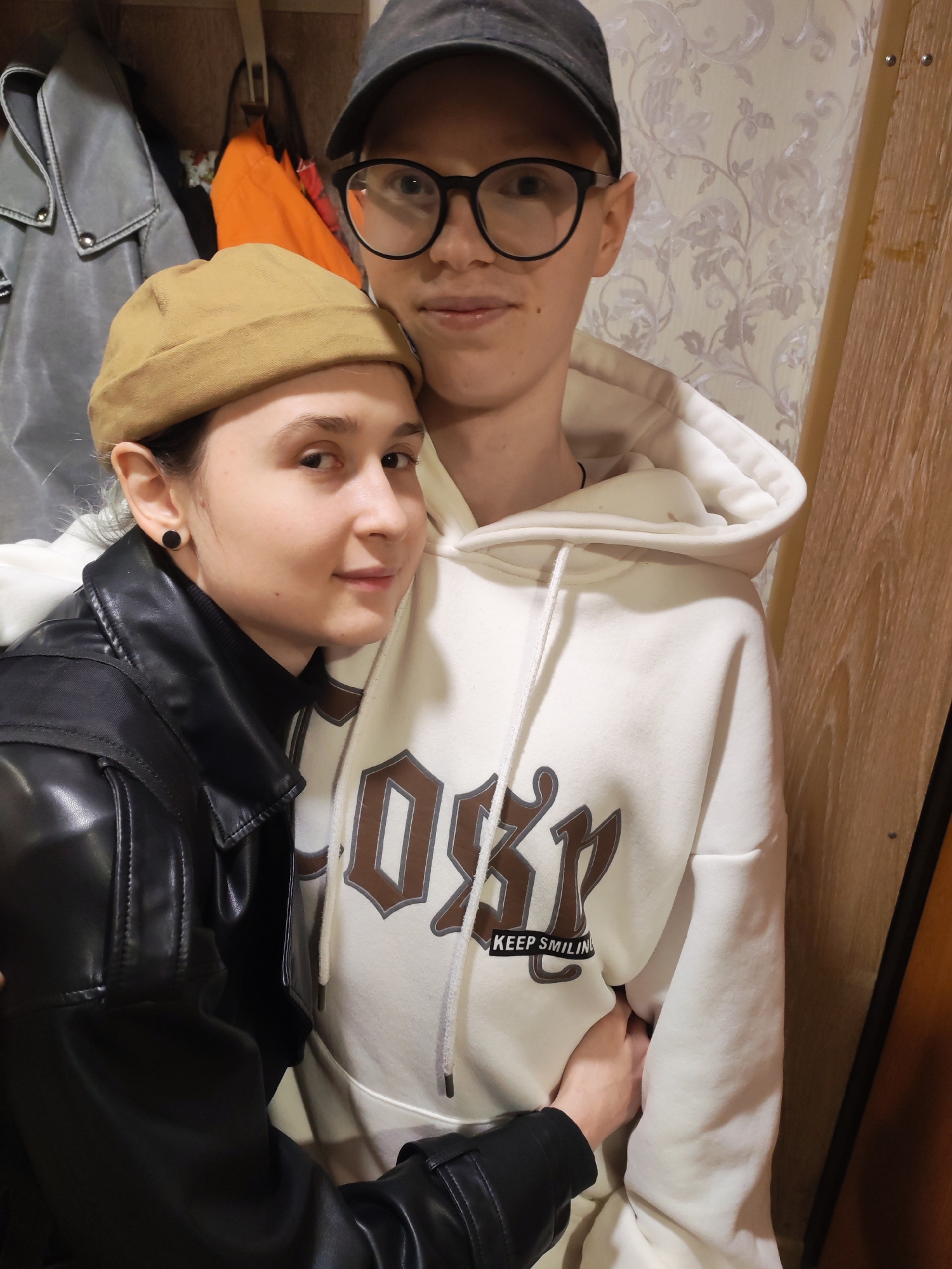 This image, taken indoors in Chaikovsky, Russia, features two individuals in what appears to be a hallway or entryway.
The person on the right, positioned slightly behind the other, wears a dark baseball cap and black-rimmed glasses, looking towards the camera with a calm expression. They are dressed in a cream-colored hooded sweatshirt featuring a large, stylized brown word design, underneath which the text "KEEP SMILING" is clearly visible. Their arm is around the waist of the person in front.
The second individual, on the left, wears a light brown docker beanie and a black leather jacket over a dark turtleneck or top. They have a small, black circular stud earring in their left ear. This person faces partly towards the camera, leaning against the first individual, with a gentle, soft expression, and their hand is seen resting on the other person's side.
The background includes a wooden wall panel on the left, from which a light grey jacket and a bright orange bag are hanging. The wall behind the person in glasses is covered with light-colored wallpaper featuring a subtle, patterned design. A vertical wooden panel frames the right side of the image. The lighting suggests an indoor setting, likely under artificial light. The overall scene conveys a sense of warmth and affection between the two subjects.