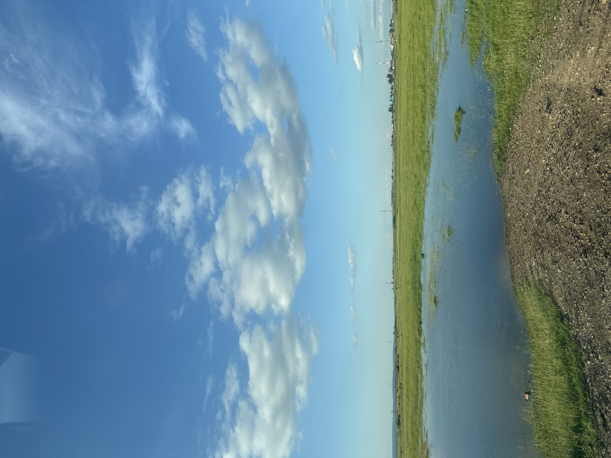 The image depicts a rural landscape under a partly cloudy blue sky. In the foreground, a body of water reflects the sky, bordered by green grass and a dirt path. The middle ground consists of a vast, flat expanse of green grass, dotted with small patches of water. In the distance, a small settlement with houses and a tall structure, possibly a communication tower, is visible on the horizon. The overall impression is one of a serene, open, and natural environment.