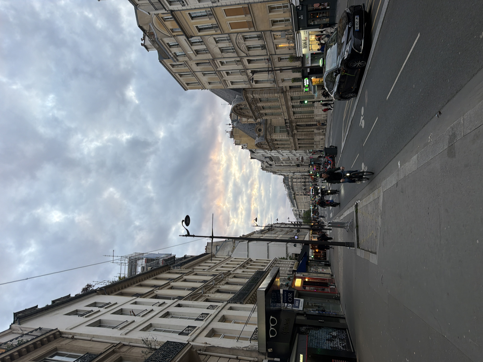 The image shows a street view in Paris, France. The street is lined with classic Parisian buildings with multiple stories and detailed facades. The sky is overcast with a mix of grey and white clouds. A street with cars parked on the right side and a designated bicycle lane is present. Several cyclists are riding down the street in the distance. Streetlights and commercial signage are also visible along the sidewalk. The overall scene is urban, likely taken during the early evening or late afternoon due to the cloudy sky and diminishing daylight. Pedestrians are walking along the sidewalks. A car is parked on the right side of the street. A store with eyeglasses in the window is shown on the bottom left.