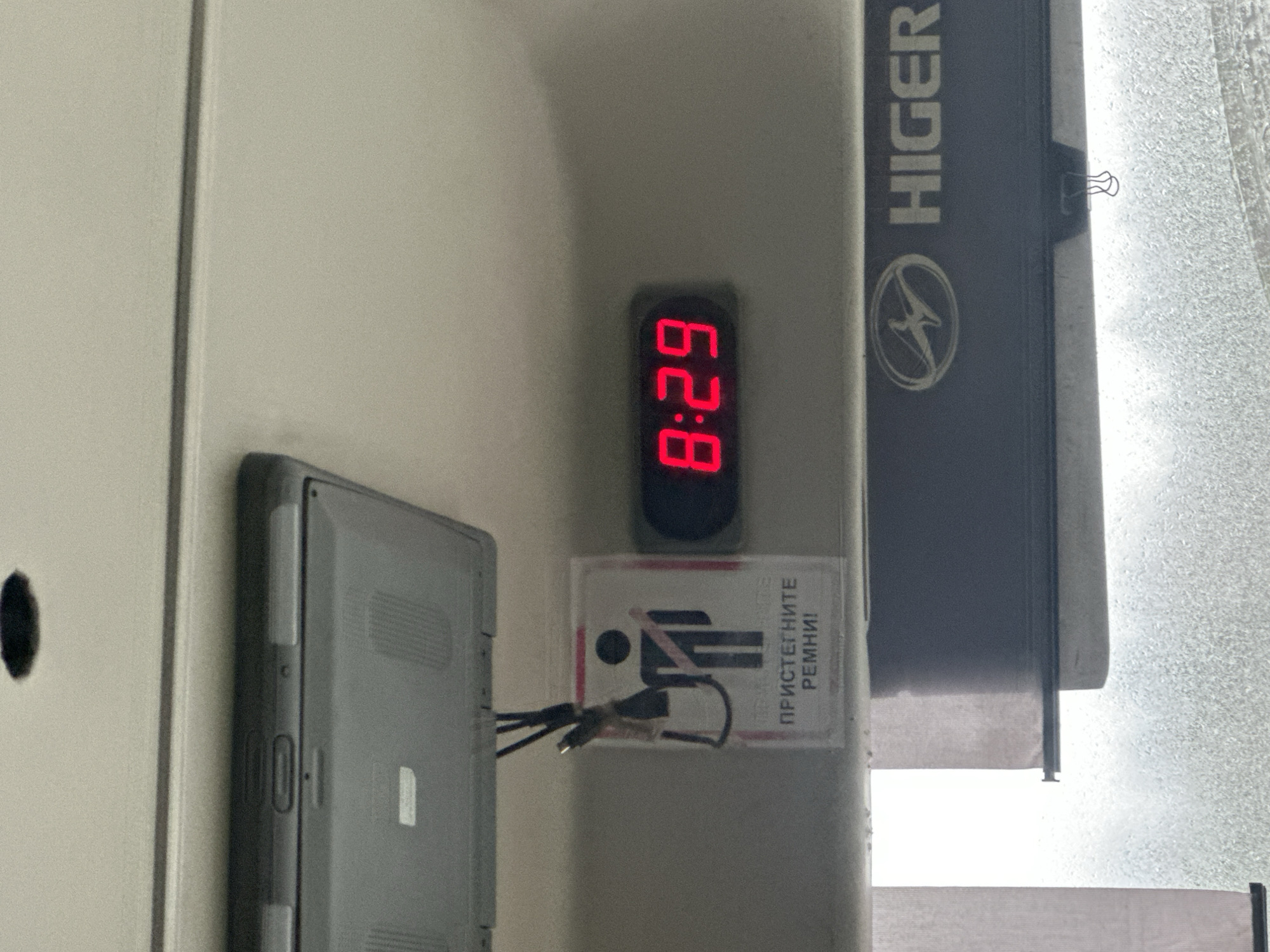 An indoor shot shows a digital clock displaying 8:29 in red digits. A sign below the clock with a graphic of a person wearing a seatbelt and Russian text "ПРИСТЕГНИТЕ РЕМНИ!" is visible. A gray laptop sits next to the clock and sign, with wires coming out from it. A "HIGER" logo is visible on the side of what appears to be a bus seat or headrest. The image might be taken inside a Higer bus. There is no information to confirm the location in the city of Krasnaya Gorka, Russia.
