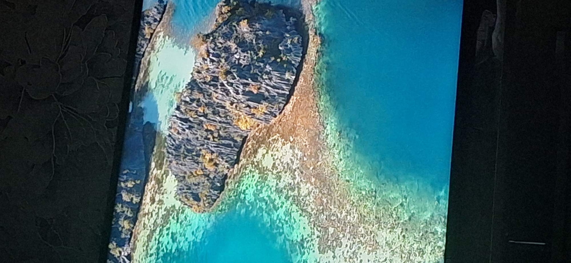 The image displays an aerial view of a rocky island amidst a clear turquoise ocean. The island is characterized by sharp, grey rock formations covered with sparse vegetation, appearing to be a karstic landscape. The water surrounding the island is shallow and exhibits a sandy or coral bottom, visible through the clear water, with areas of varying light and dark patches indicating depth or submerged features. The overall scene is one of a tropical or subtropical marine environment. There is no visible indication of the city of Bila Tserkva, Ukraine, or any urban elements within the frame.