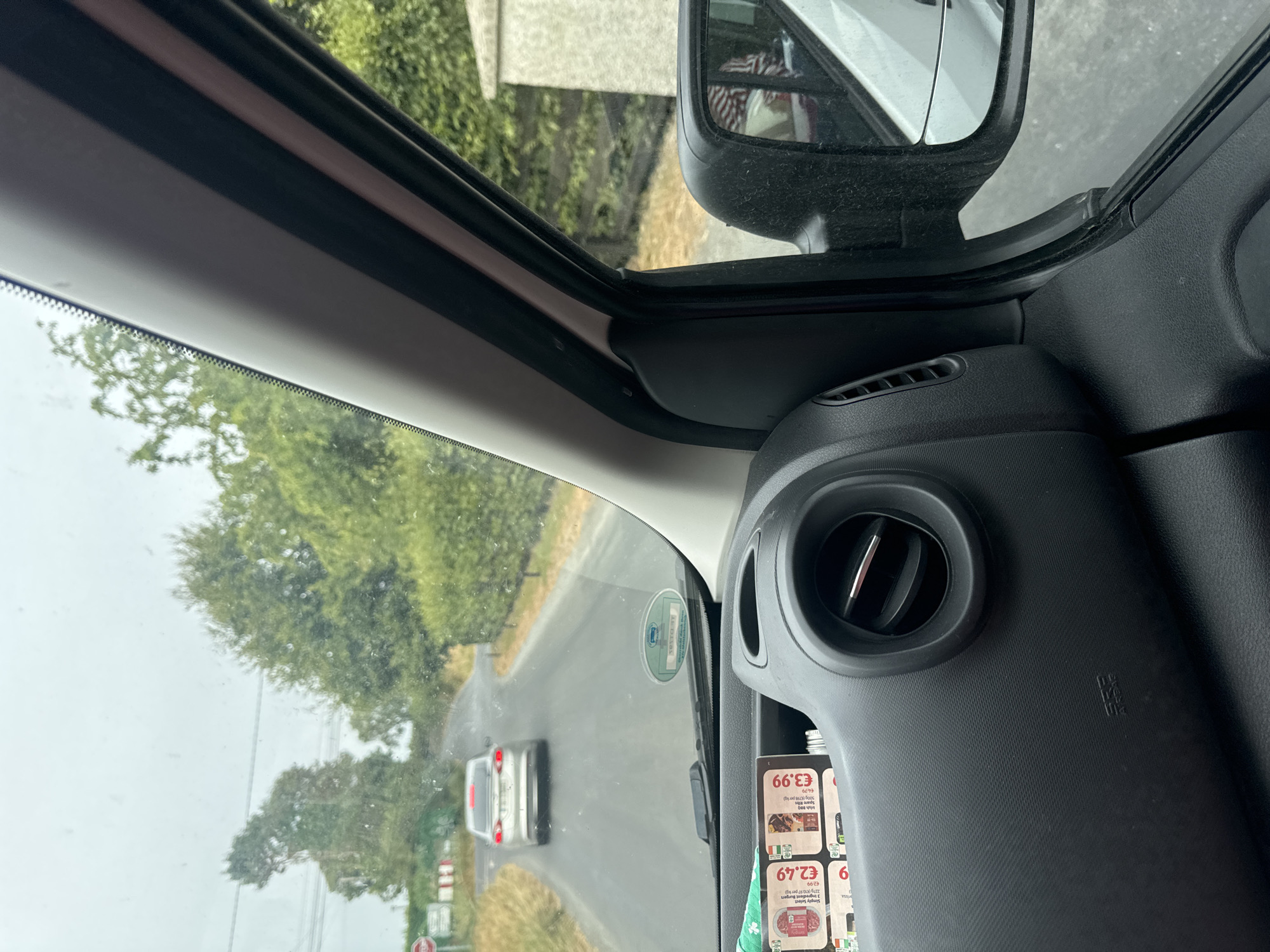 An external, slightly hazy, eye-level, medium shot captures a view from inside a vehicle looking out at a two-lane asphalt road. The road curves to the left and is lined with green foliage and trees. A silver sedan is driving away from the camera on the road. The sky is overcast and grey. On the dashboard in the foreground, there are two product advertisements with prices displayed, an air vent, and an airbag warning label. The side mirror of the vehicle is visible on the right, reflecting a red and white patterned object, likely clothing. The image appears to be taken in Bamford Cross, Ireland, based on the context provided.